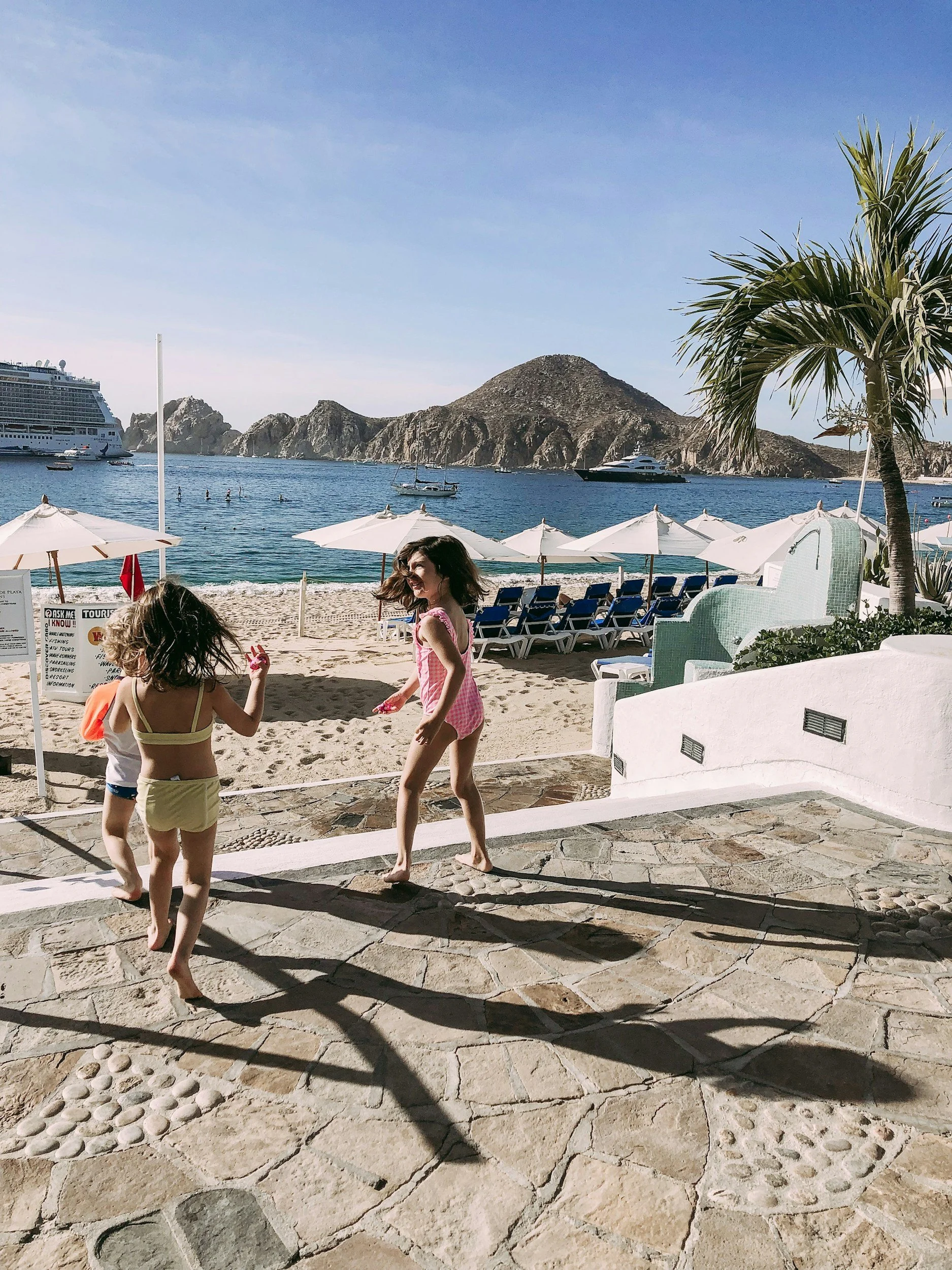 Two children playing on a beach with umbrellas, chairs, and a view of boats and mountains in the background.
