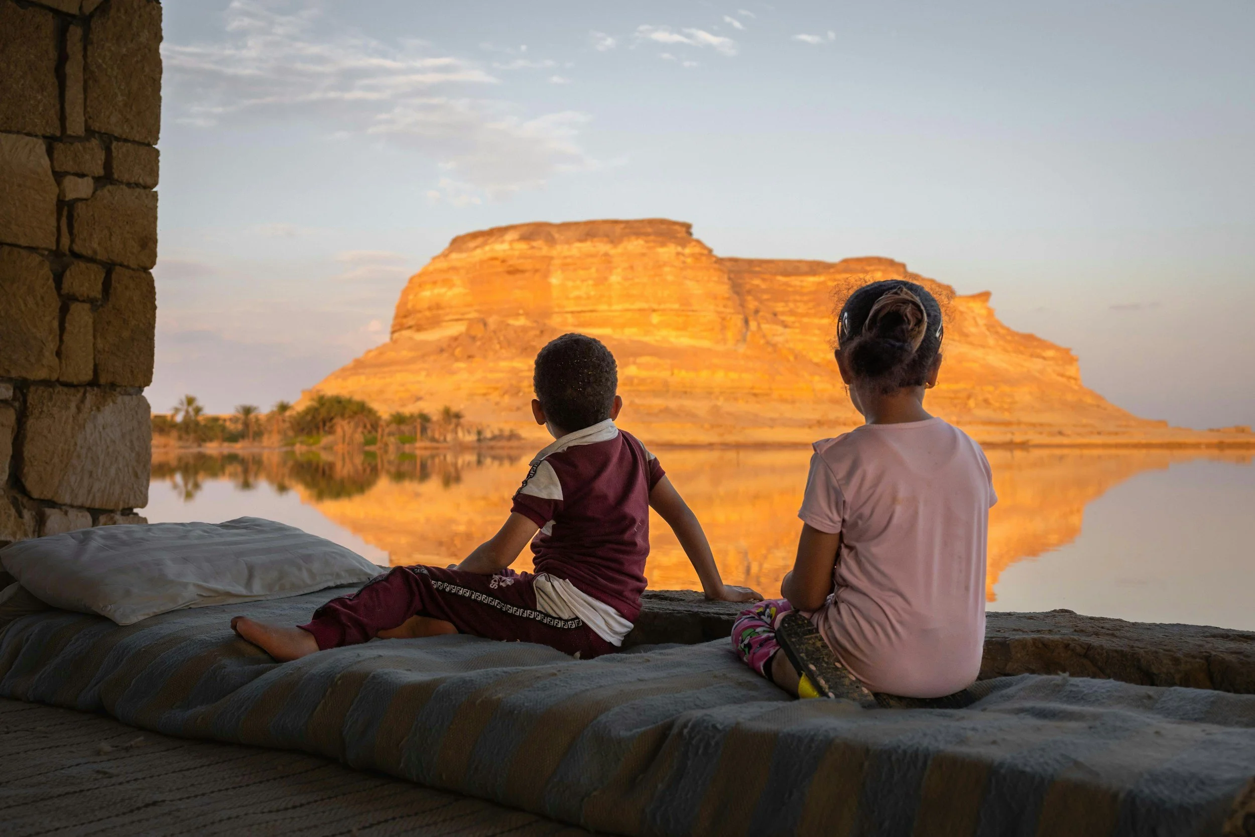 Two children sitting on bedding in an outdoor setting, looking at a large, illuminated rock formation across a body of water during sunset.