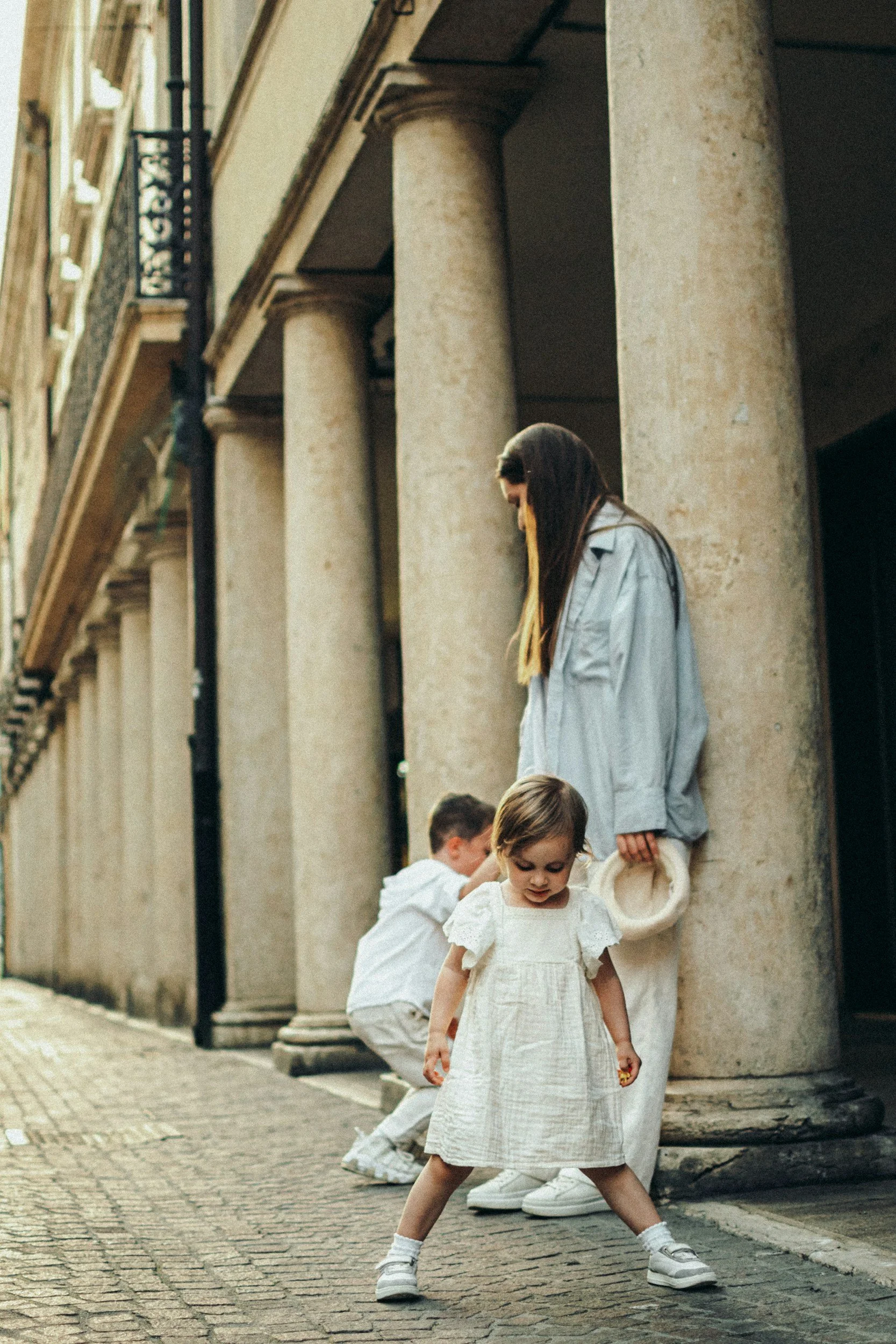 Children playing outdoors on a cobblestone street, with an older woman and historic building columns in the background.