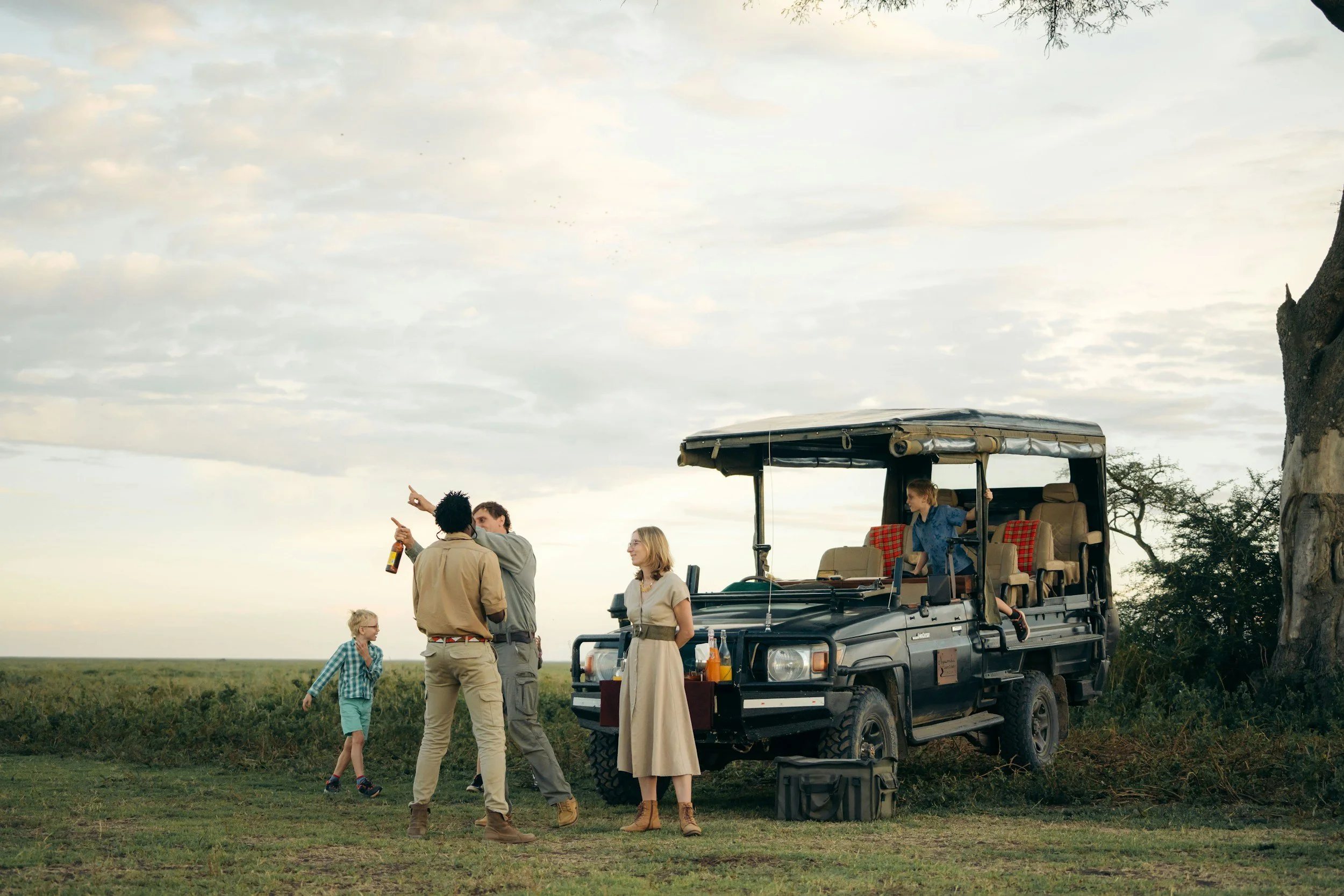 Group of people talking near an open safari vehicle in an open field with a tree and cloudy sky in the background.