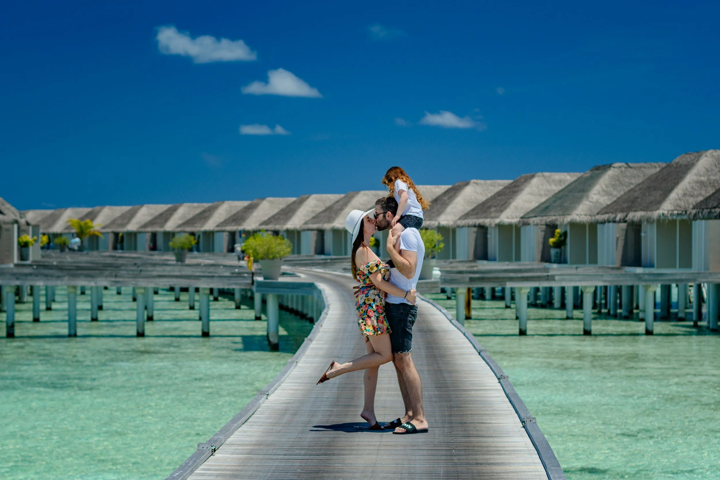 A couple kissing on a wooden dock over clear green water, with a young girl on the man's shoulders, all under a blue sky with a few clouds. The dock leads to overwater bungalows with thatched roofs.
