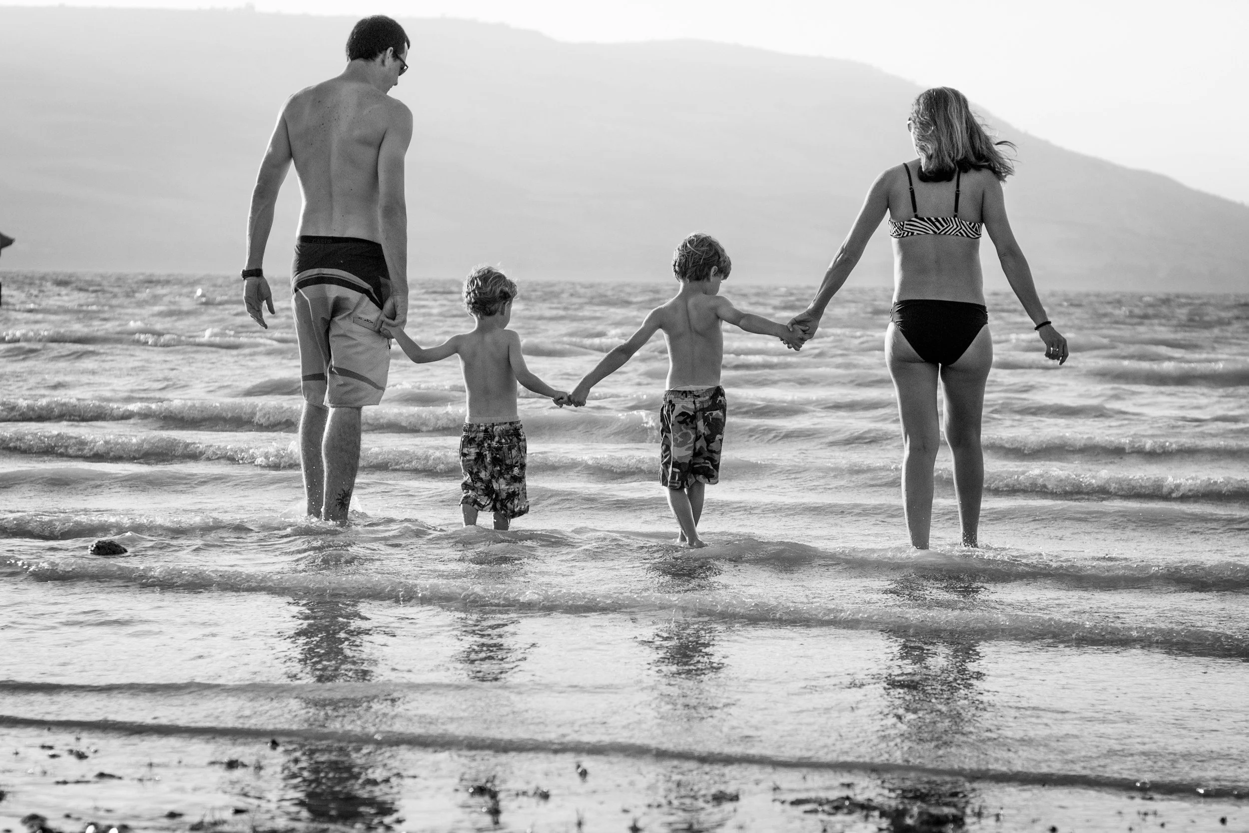 A black-and-white photograph of a family of five walking along the shoreline at the beach, holding hands. The family includes an adult male, an adult female, and three young children. The background features a hilly landscape and the ocean.
