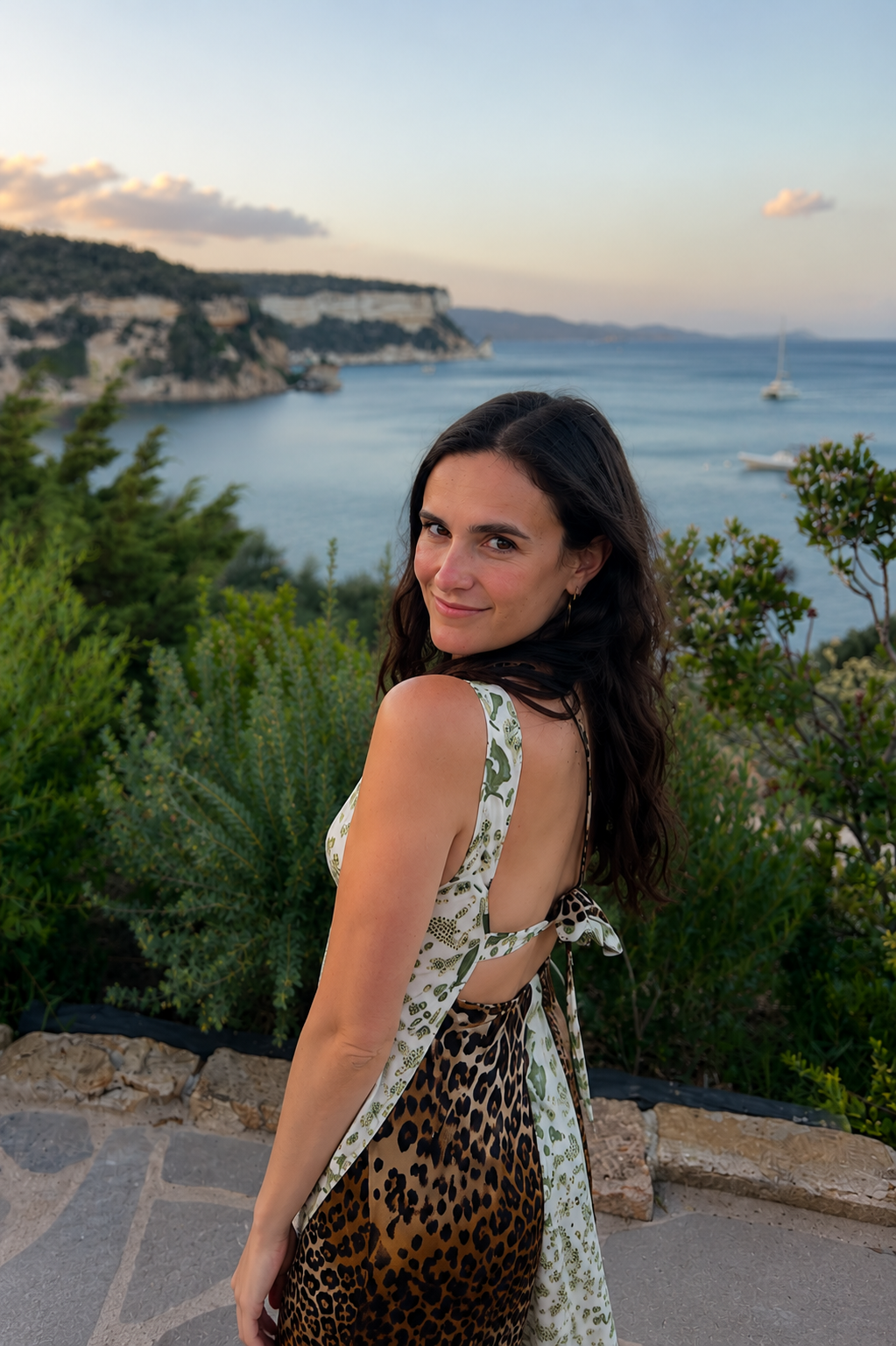 A woman with dark wavy hair, smiling, standing on a stone path with greenery, overlooking a body of water with sailboats and cliffs in the distance during sunset.