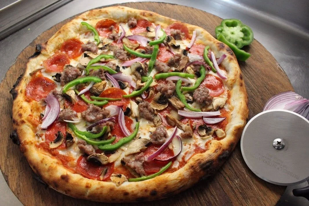 Pizza con pepperoni, carne molida, cebolla, pimiento verde y champiñones, servida en tabla de madera.