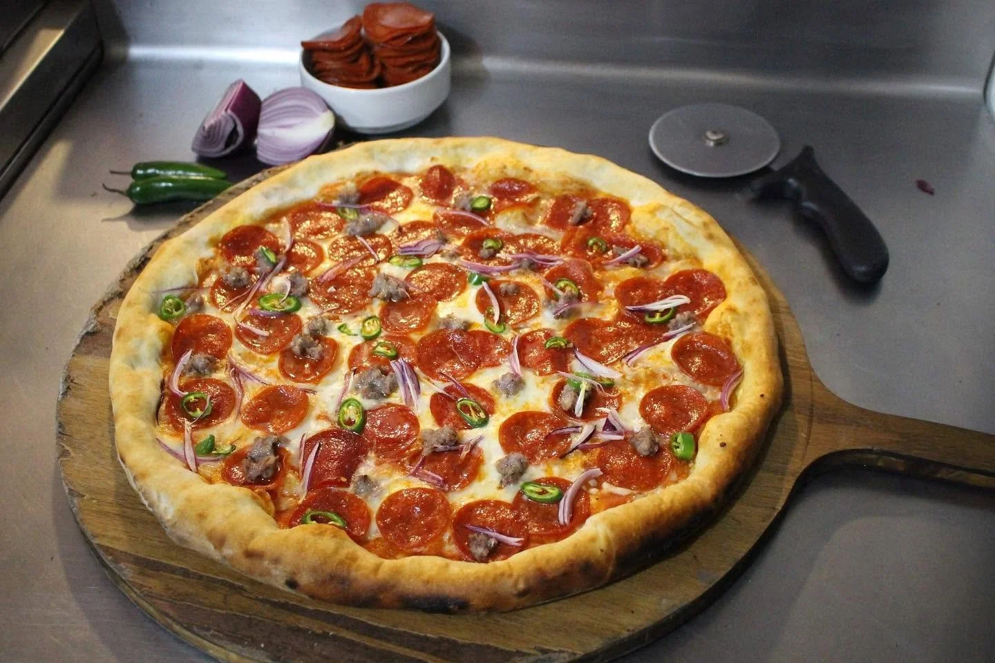 Pizza con pepperoni, cebolla morada, chiles verdes y carne en una tabla de madera en una cocina.