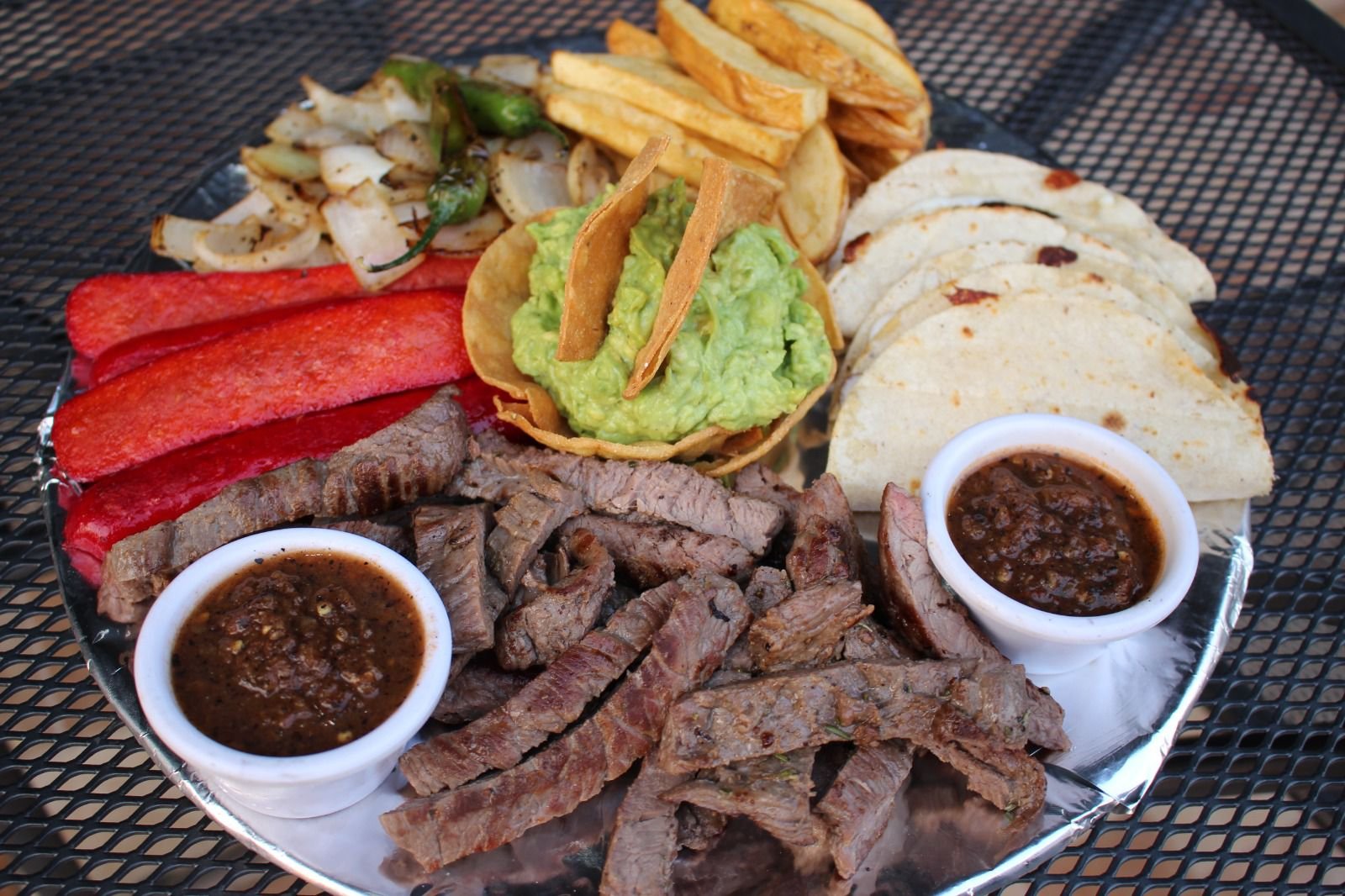 Plato de comida mexicana con carne asada, guacamole, salsa, frijoles, tortillas, papas fritas, cebolla asada, y pimientos. Incluye salsas y tortillas en un plato de metal.