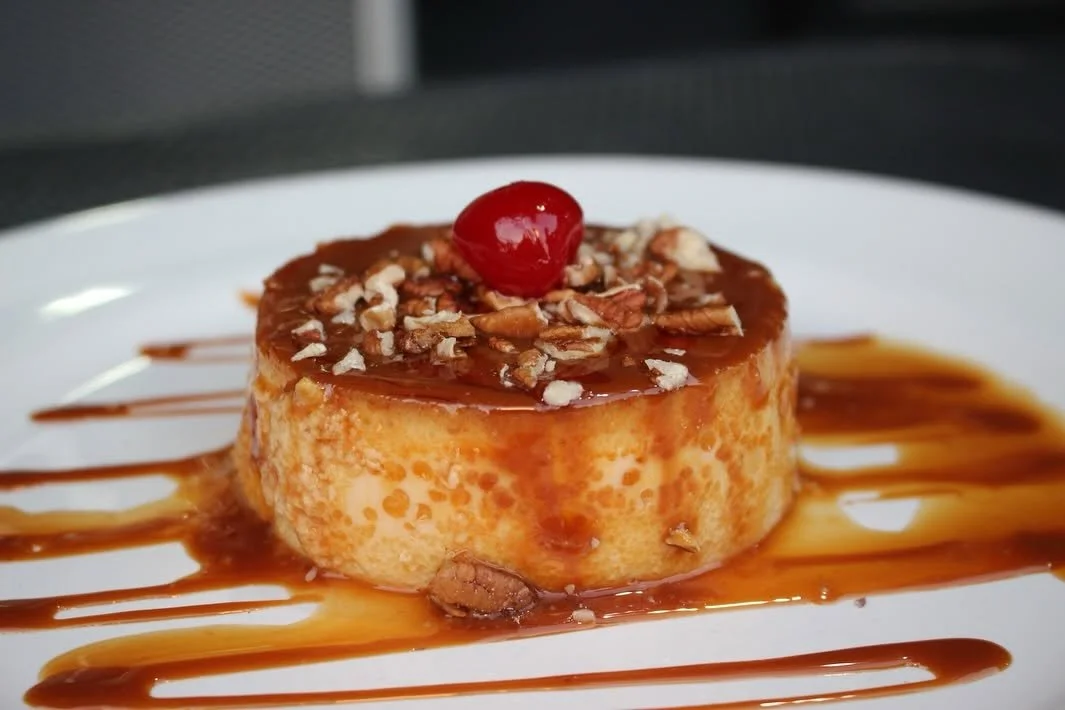 Postre tipo flan con gluten de caramelo, decorado con cereza y toppings de nueces en un plato blanco.