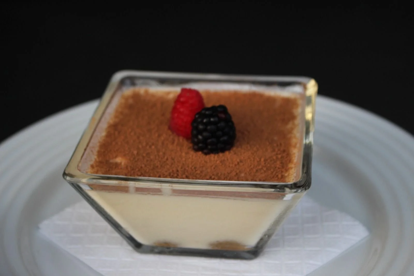 Postre de tiramisú en un vaso cuadrado con frambuesa y mora encima, sobre un plato blanco.