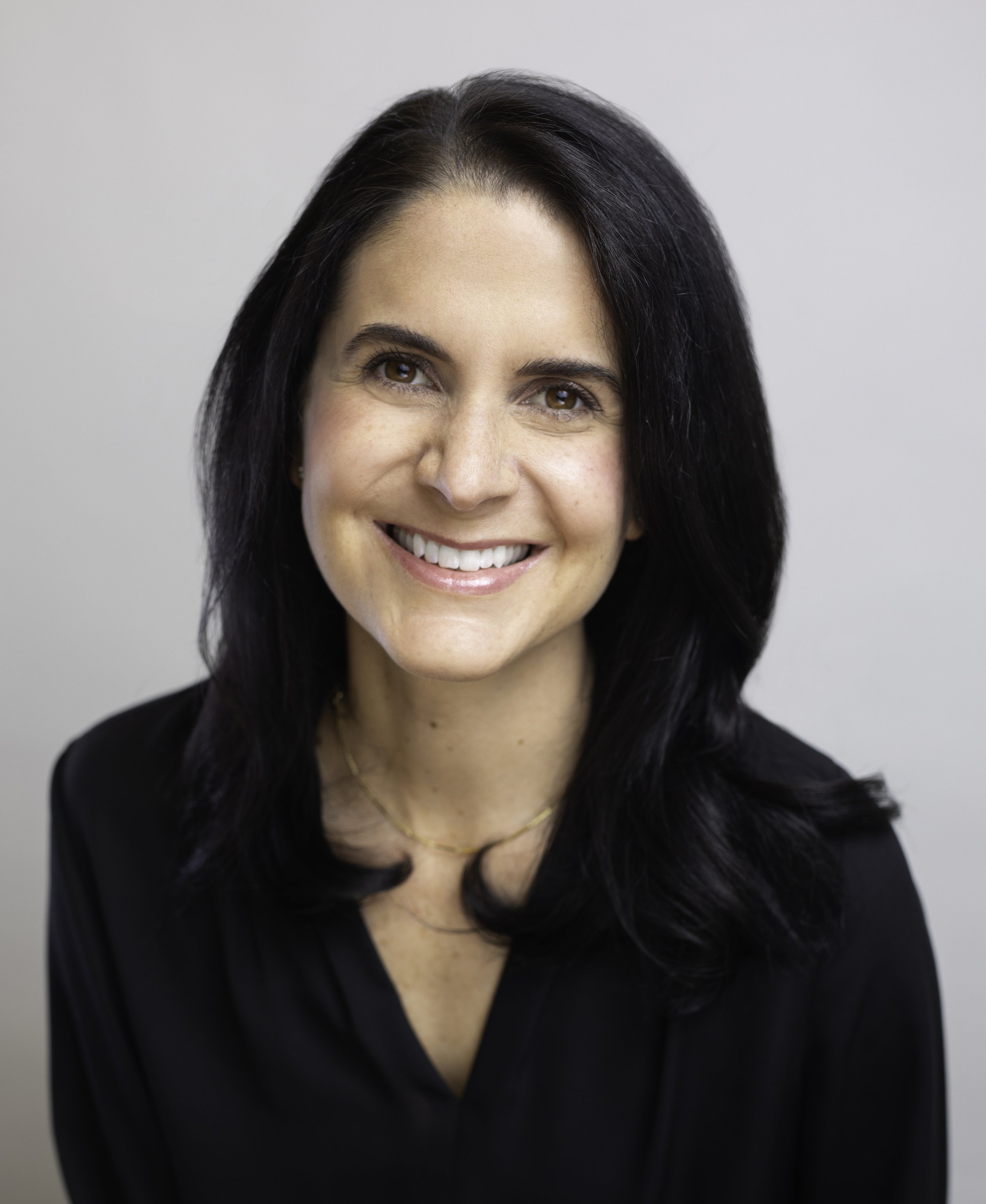 Close-up of a smiling woman with dark hair, wearing a black top and a gold necklace, against a plain light background.