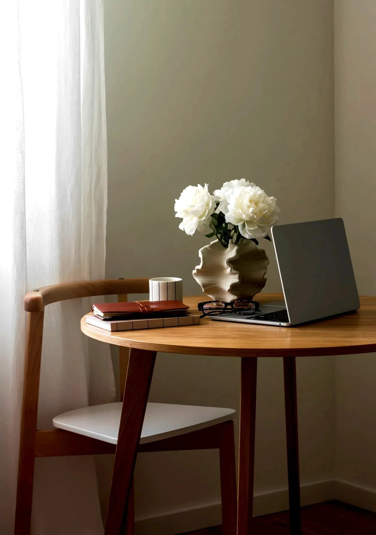 A cozy wooden desk with a plant in a white vase, a laptop, glasses, and notebooks in a softly lit room with a curtain and plain wall.