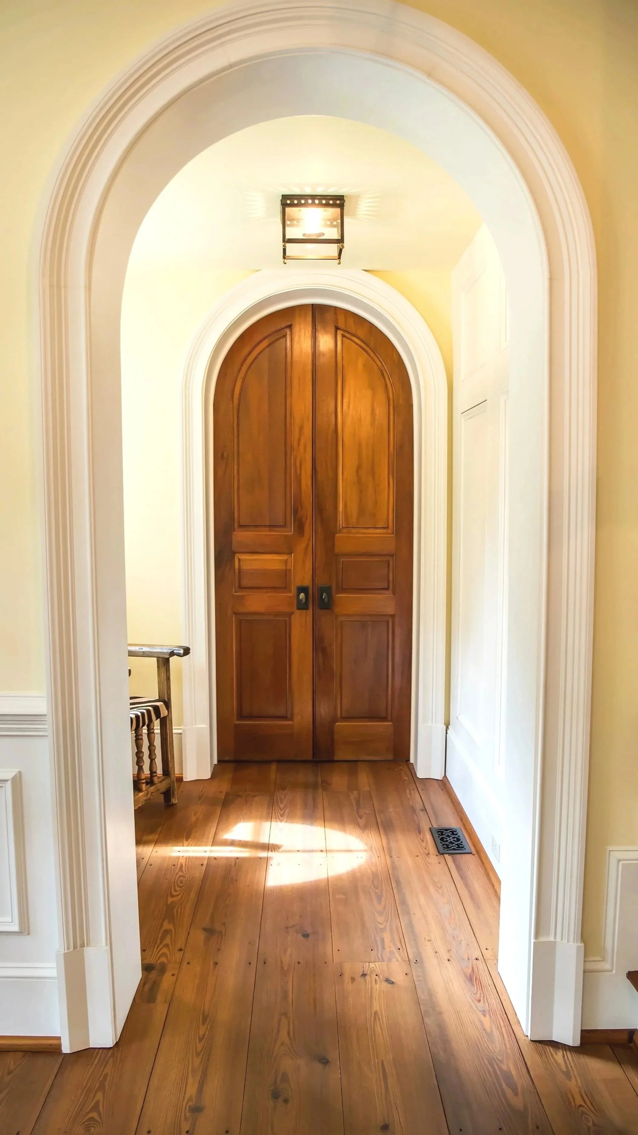 Wooden double door in an arched doorway with white trim, yellow walls, hardwood floor, and a wall vent on the floor to the right.