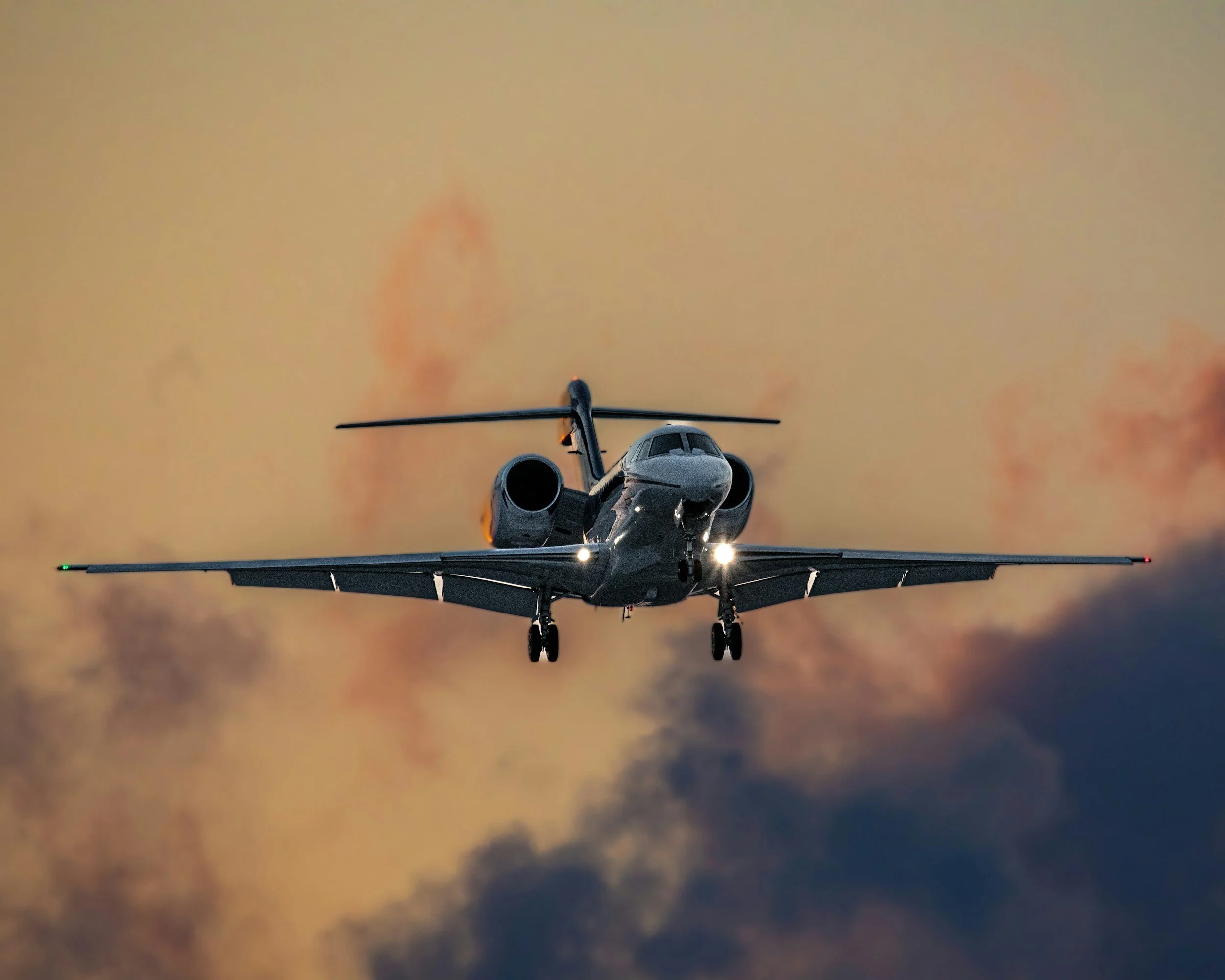 A private jet flying through the sky during sunset or sunrise, with an orange glow in the background.