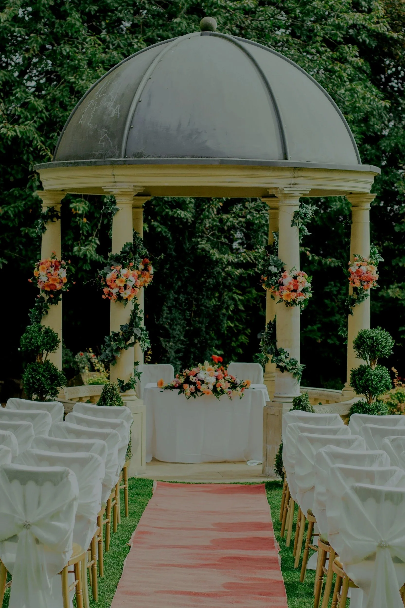 An outdoor wedding setup with white chairs and decorated with pink fabric bows, facing a decorated altar with flowers under a domed pavilion supported by columns with greenery in the background.