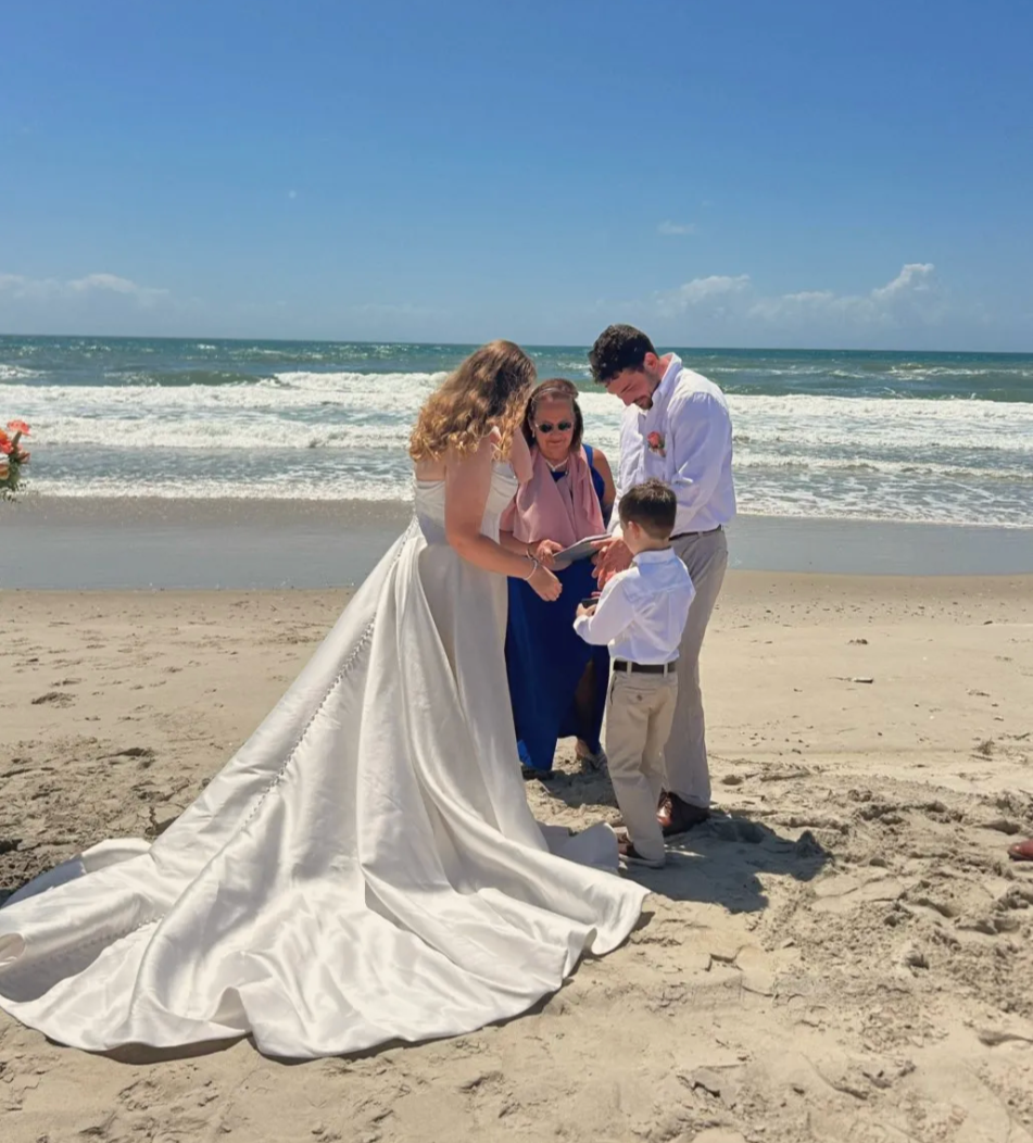A wedding ceremony on the beach. The bride is in a white gown and the groom is in a white shirt and a young boy brings the rings to  the officiant, Maritsa Hartlove.