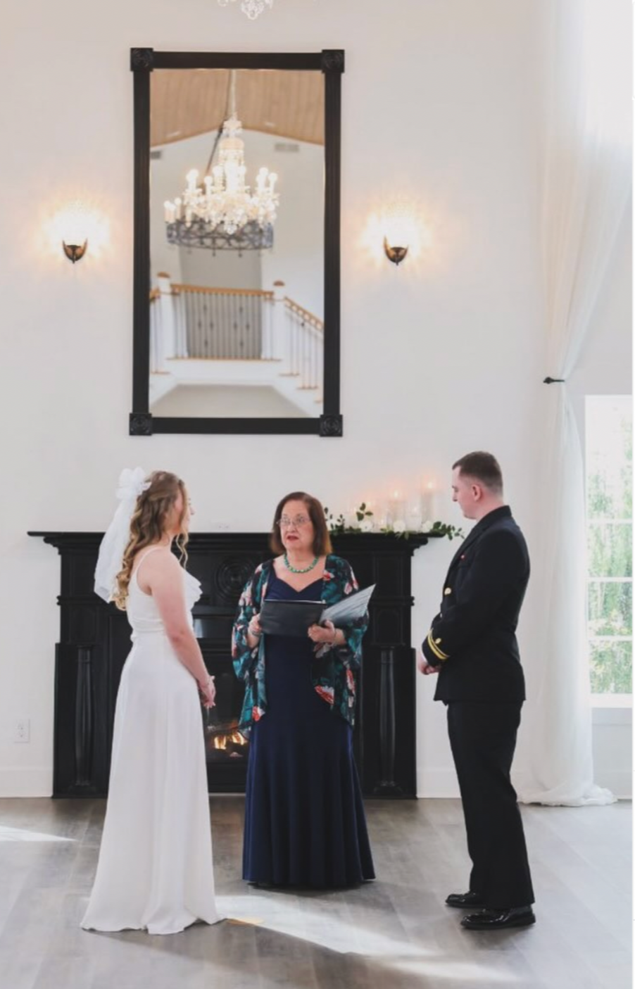A bride and groom standing before the officiant, Maritsa Hartlove, during their wedding ceremony in a bright, elegant room with high ceilings, a large mirror and a chandelier.
