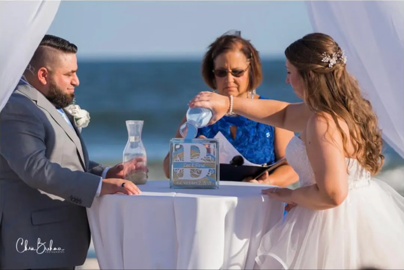 A couple getting married on a beach, their officiant holding a book, with the bride pouring water into a glass during the ceremony, all under a white canopy with the ocean in the background.