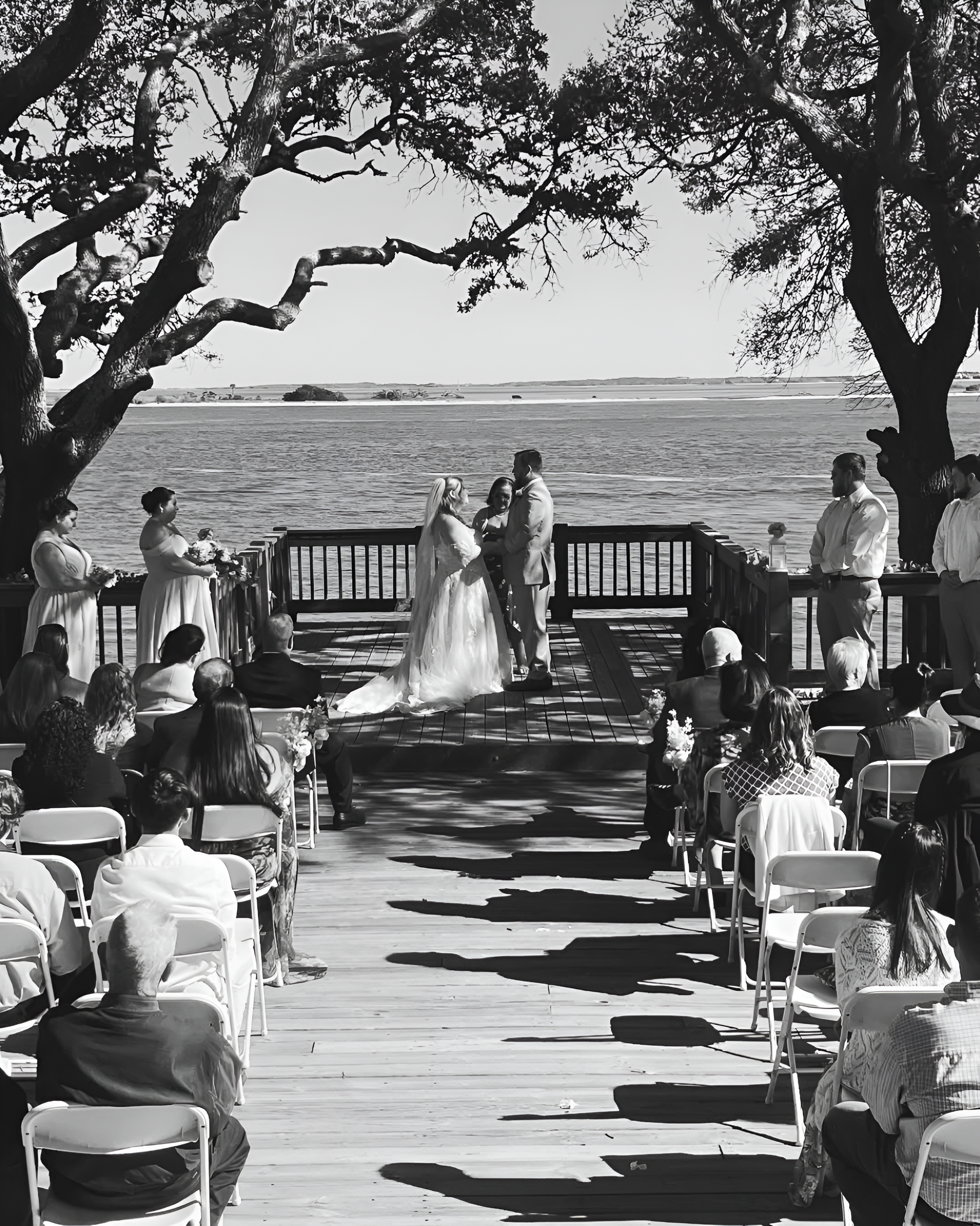 A black and white photo of a wedding ceremony on a deck by the water. The bride and groom stand under a tree surrounded by guests seated in chairs while Maritsa Hartlove officiates the wedding.
