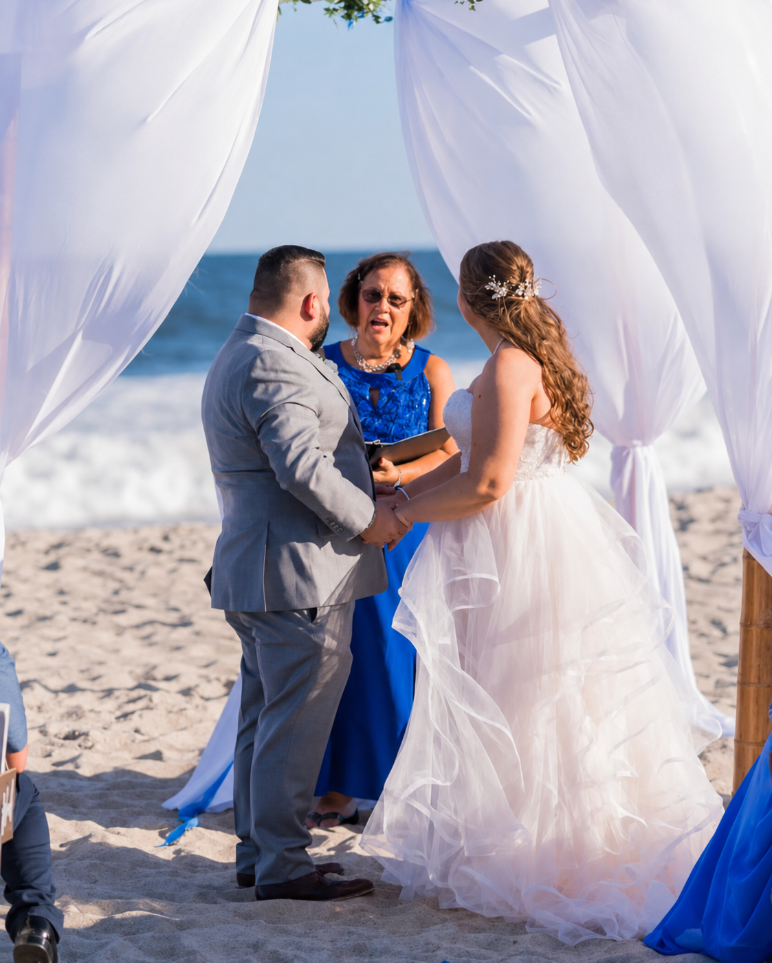 A beach wedding with a couple holding hands and facing the officiant, Maritsa Hartlove, under a white fabric canopy with the ocean in the background.