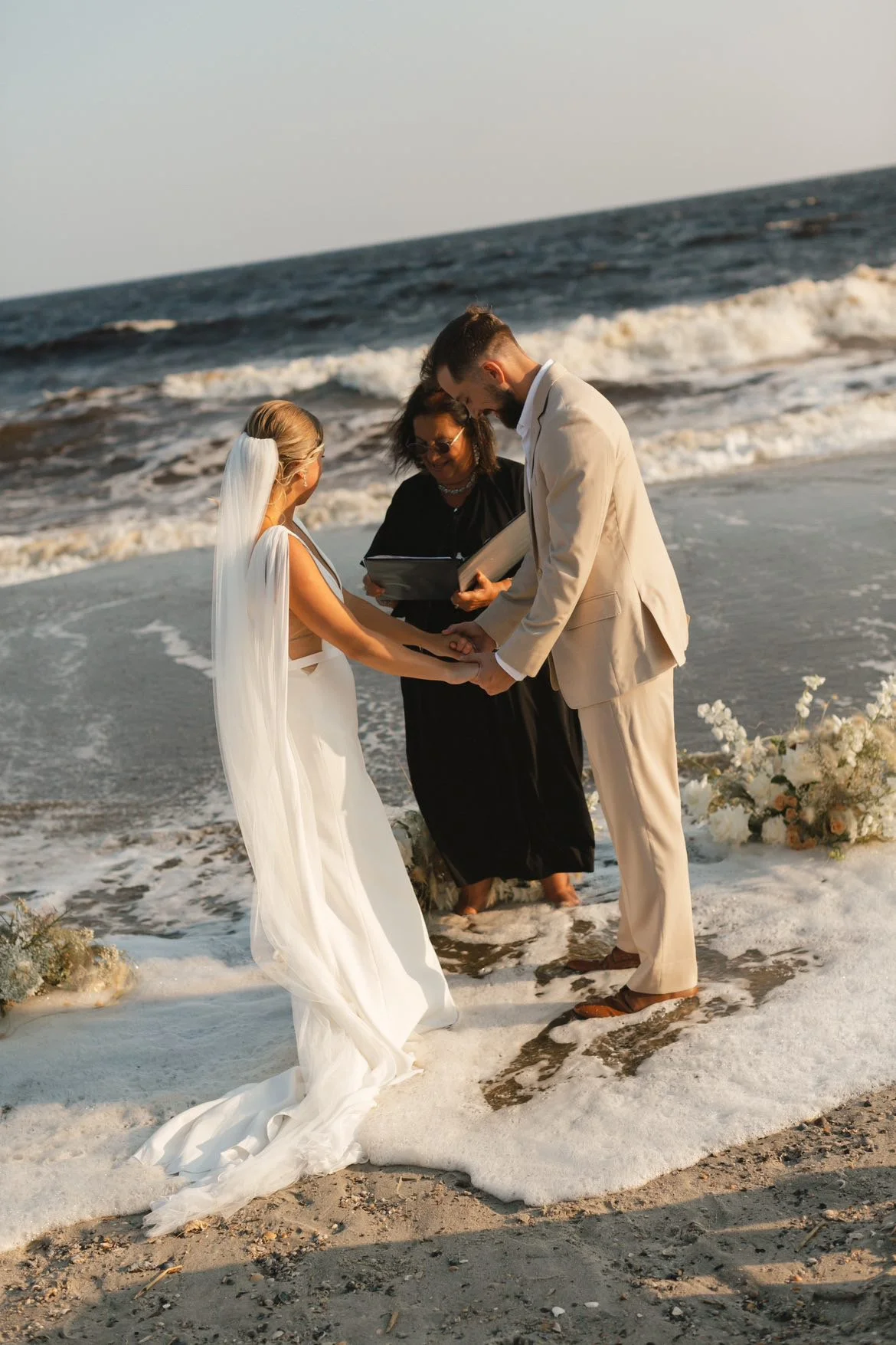 A couple getting married on the beach with the wedding officiant, Maritsa Hartlove, holding hands and standing in the sand.