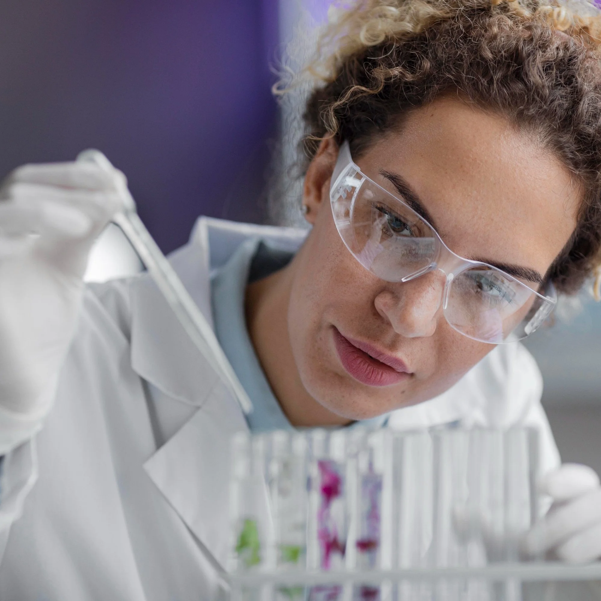 Close-up of scientist wearing safety glasses examining test tubes with colorful liquids in a laboratory.