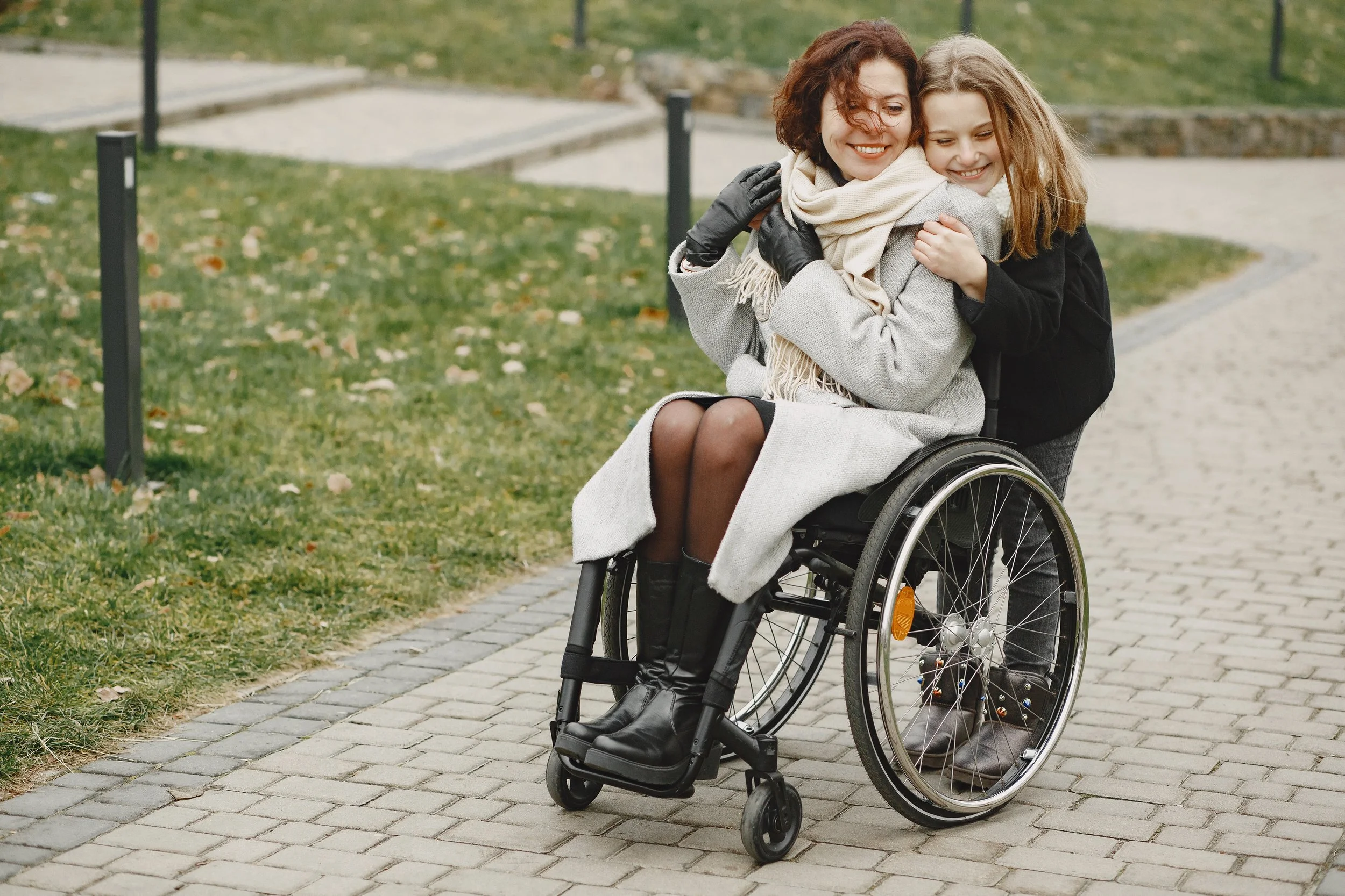 Two women are hugging, one sitting in a wheelchair, outdoors on a paved path with grass and fallen leaves.