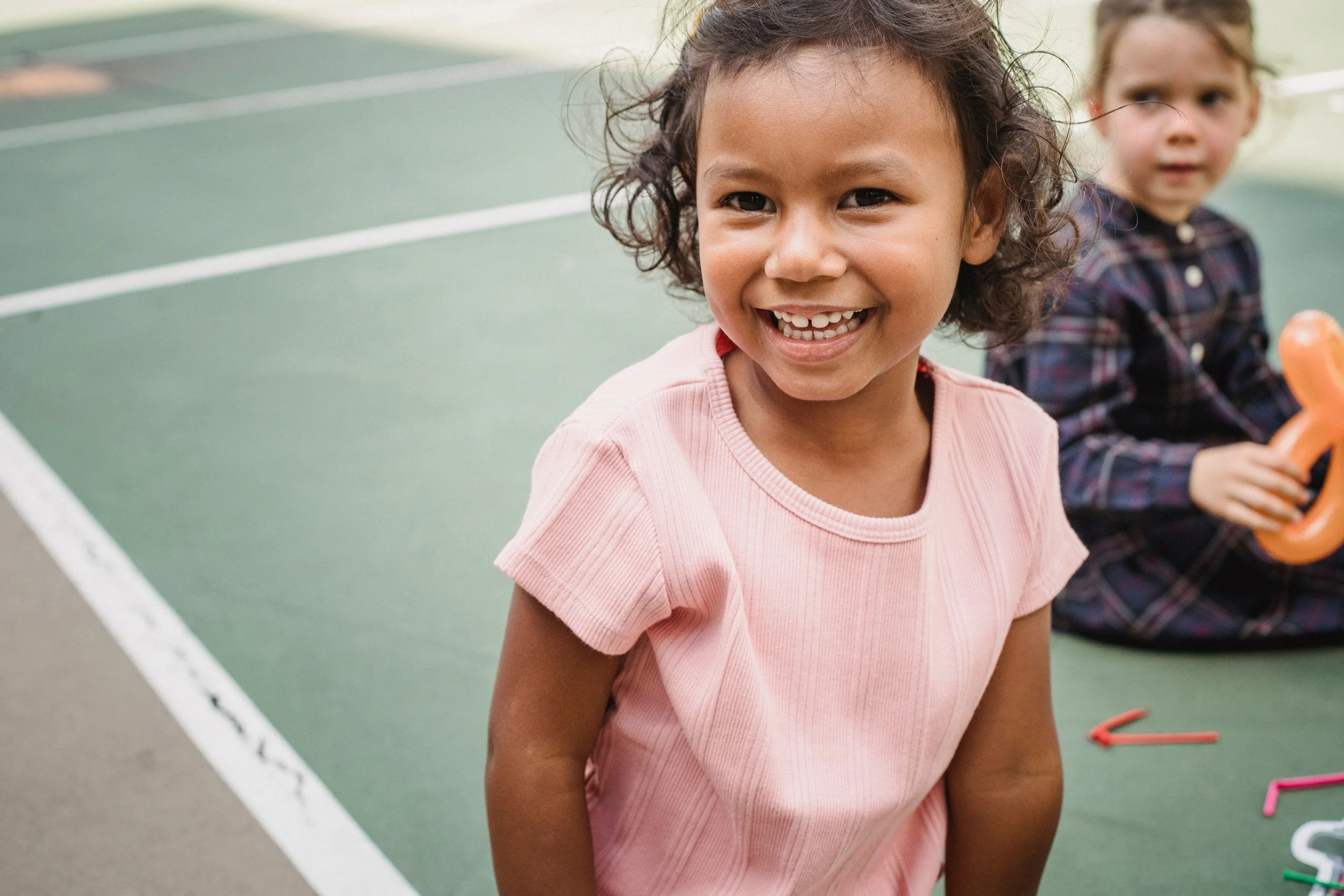 Two young girls playing outside on a tennis court. The girl in the foreground has curly hair, is smiling, and is wearing a pink shirt. The girl in the background has straight hair, is looking to the side, and is wearing a plaid shirt. Colorful chalk pieces are on the ground.