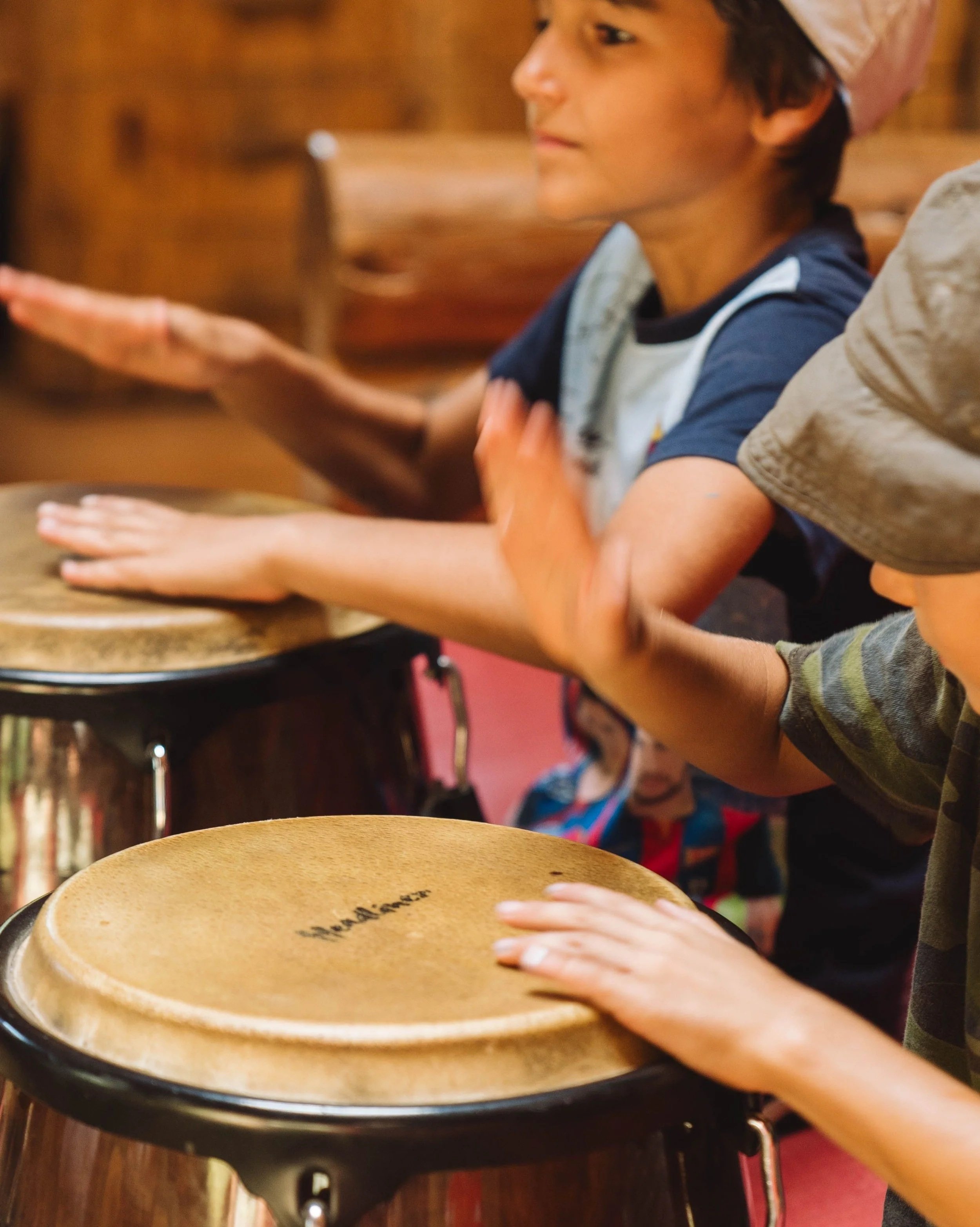 A young boy with dark hair wearing a chef's hat and dark shirt, reaching out to touch a drum in front of him, inside a cozy room with wooden furniture and a red wall.