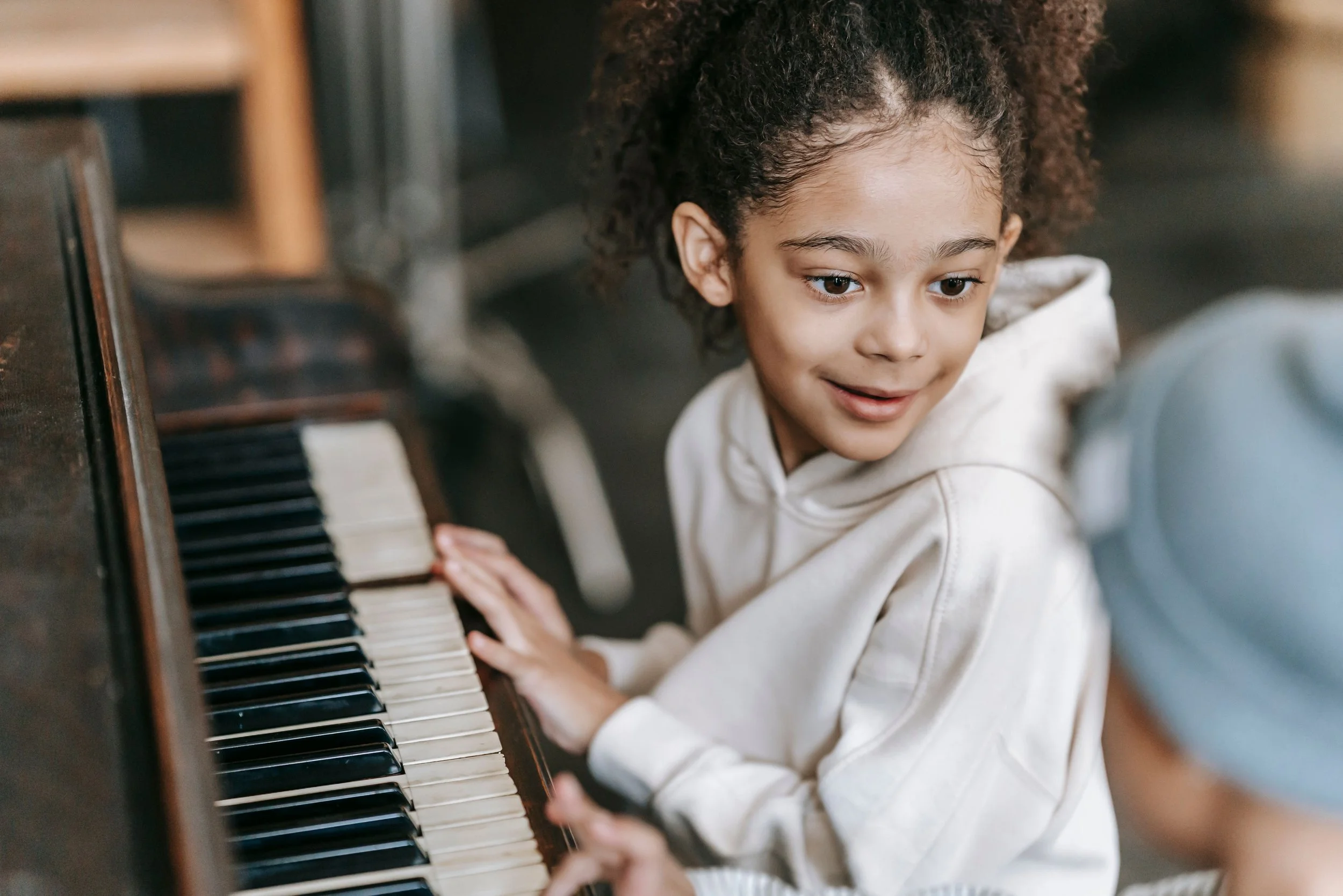 A young girl with curly hair playing a piano, looking down at the keys with a smile.