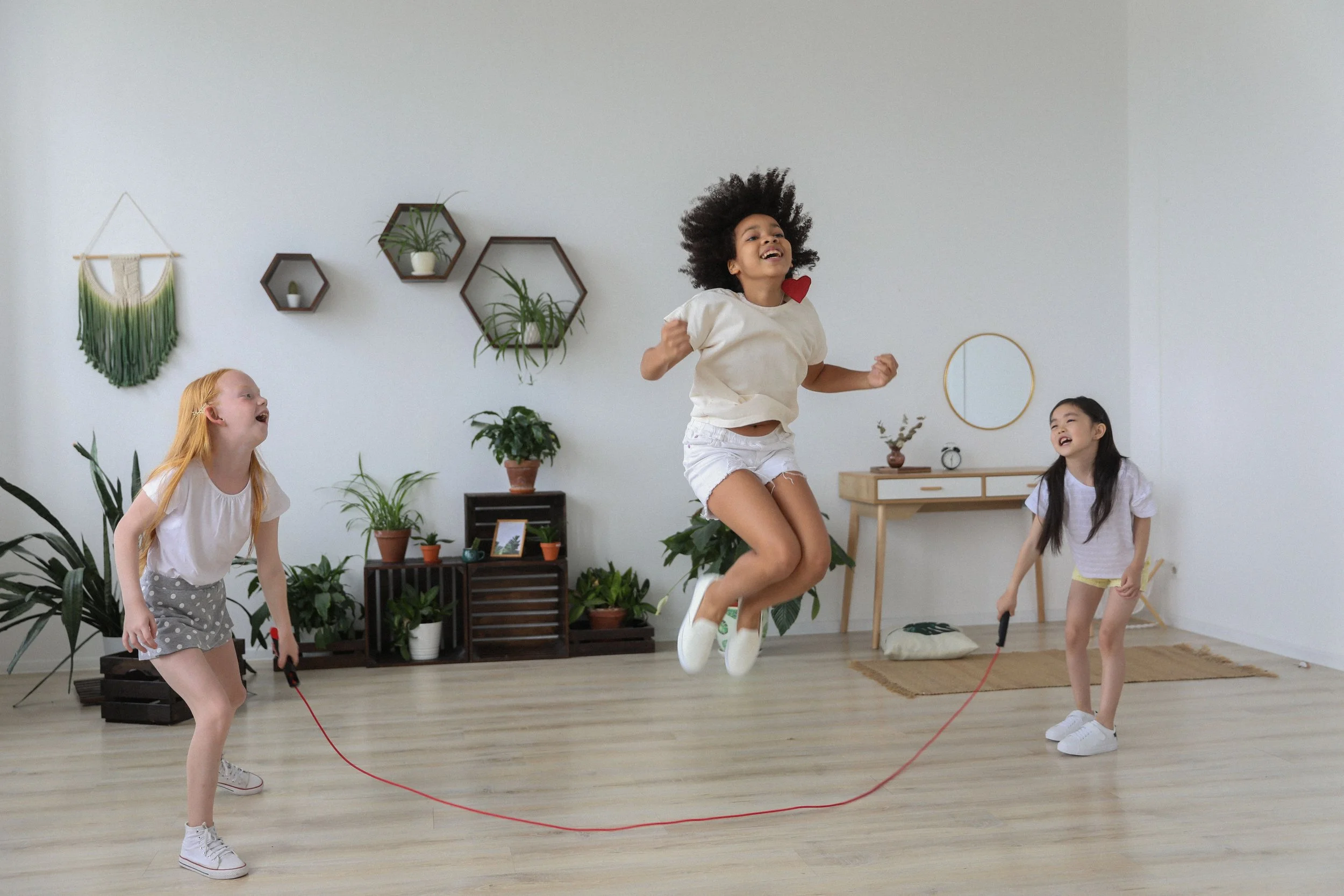 Four children are playing indoors, with one girl jumping rope and the other three children watching and laughing.