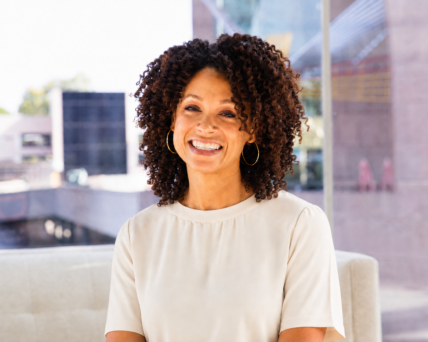 A smiling woman with curly hair wearing a cream-colored top and gold hoop earrings, sitting indoors near a window.