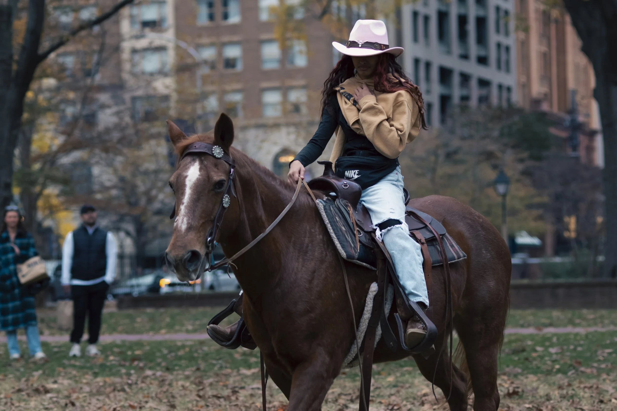 A woman riding a brown horse in a city park, wearing a tan coat and a pink hat, with buildings and people in the background during autumn.
