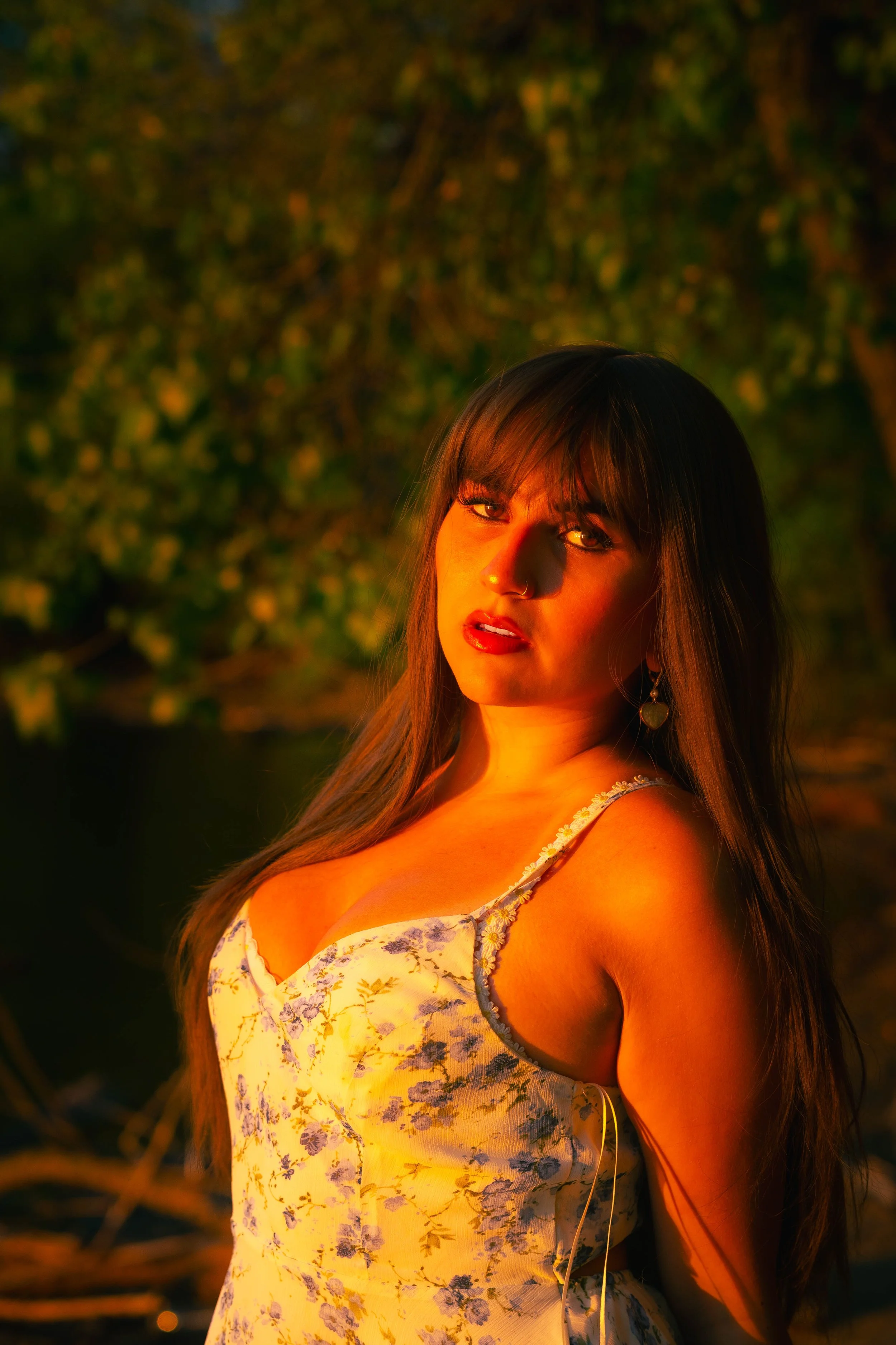 A woman with long brown hair looking at the camera, with warm lighting illuminating her face and a background of blurred greenery and trees.