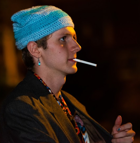 Person wearing a blue knitted beanie with a white stripe, cigarette in mouth, wearing earrings, dark jacket, and a patterned shirt, holding a cigarette in hand, with a dark background.
