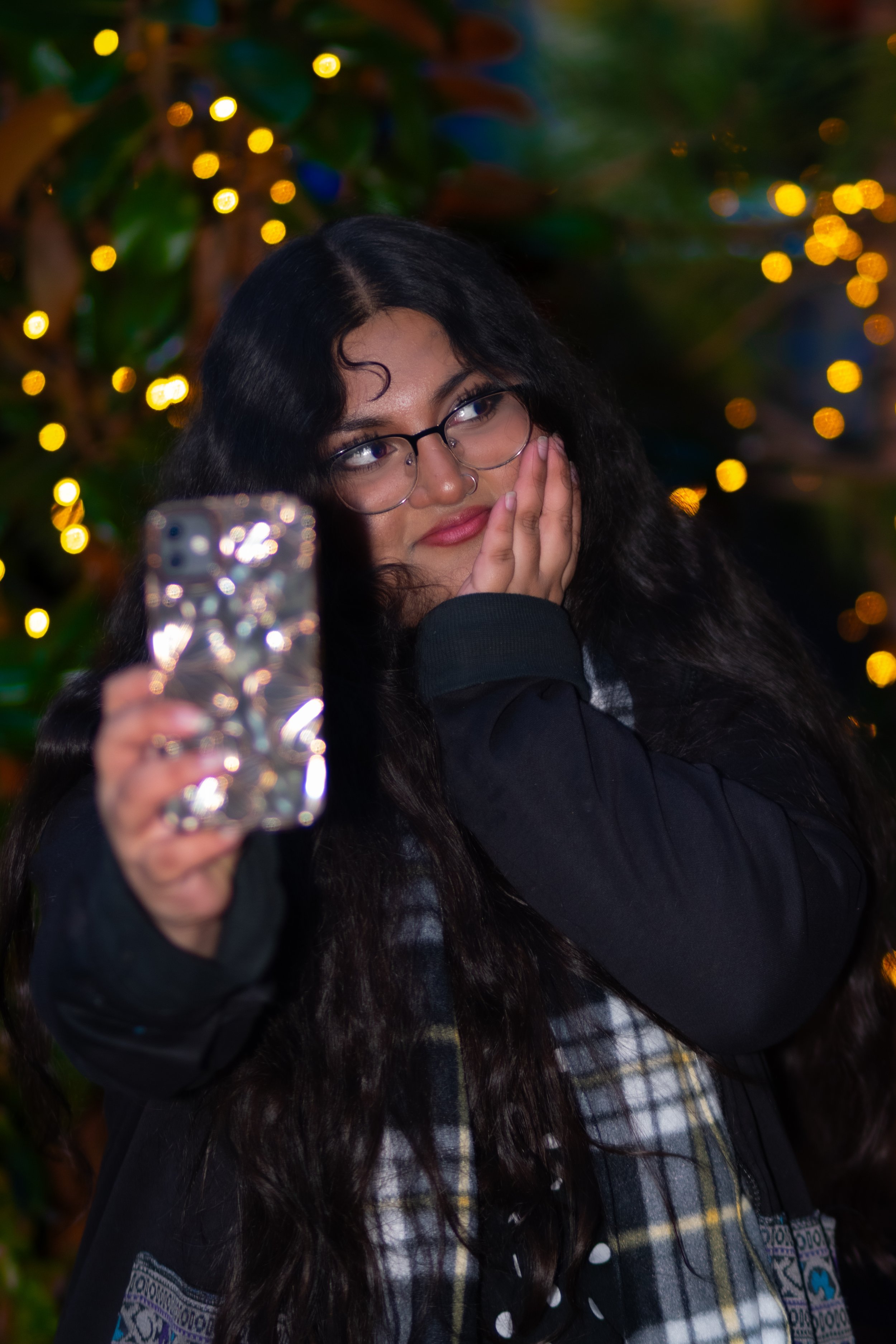 A young woman with glasses, long dark curly hair, and a nose ring is taking a selfie with her smartphone, which is visible in the foreground. She is wearing a dark jacket and a plaid shirt underneath. The background is decorated with warm, blurred string lights and green foliage.