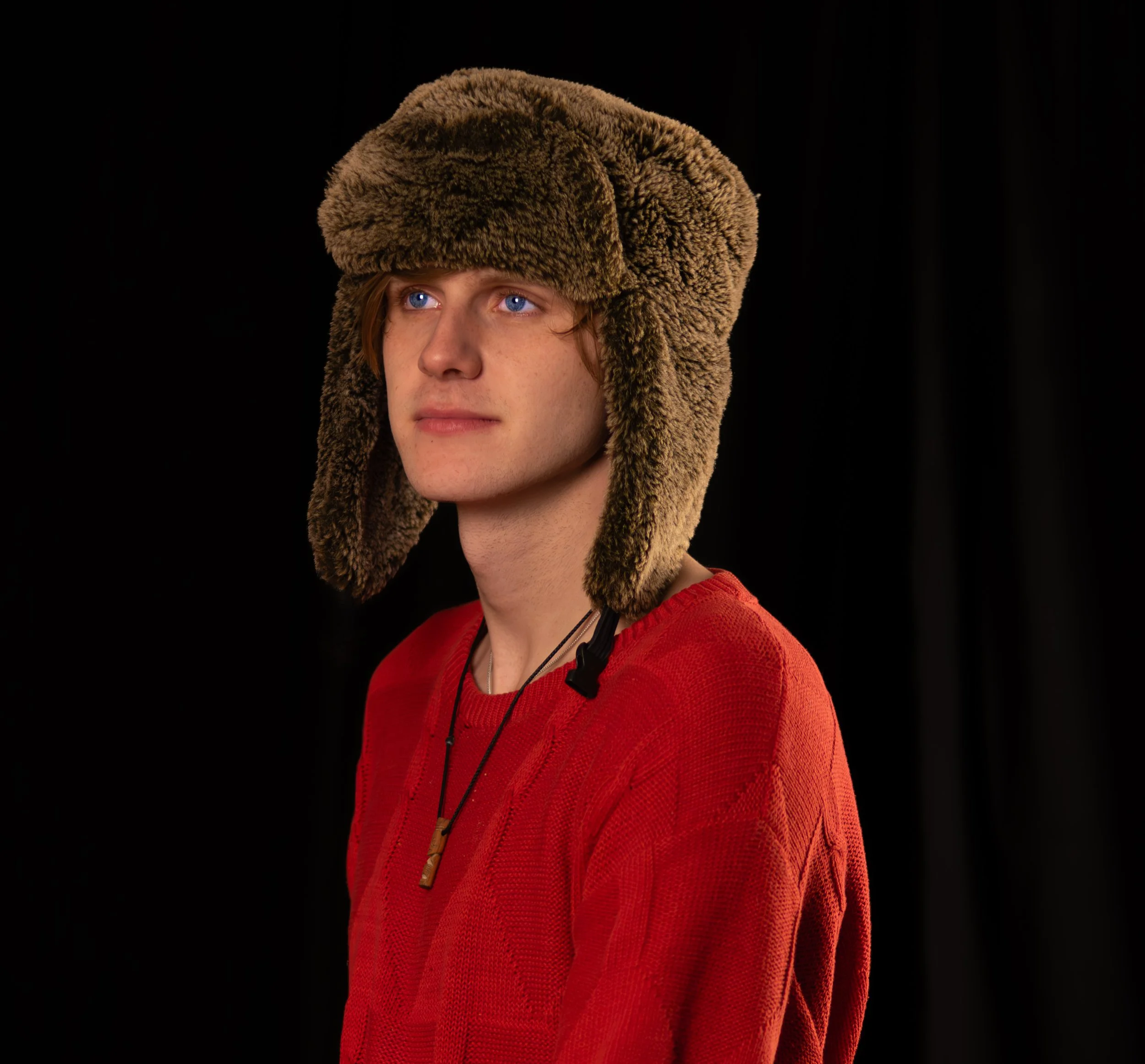 A person with light skin and blue eyes wearing a brown furry winter hat with ear flaps and a red knitted sweater, against a black background.