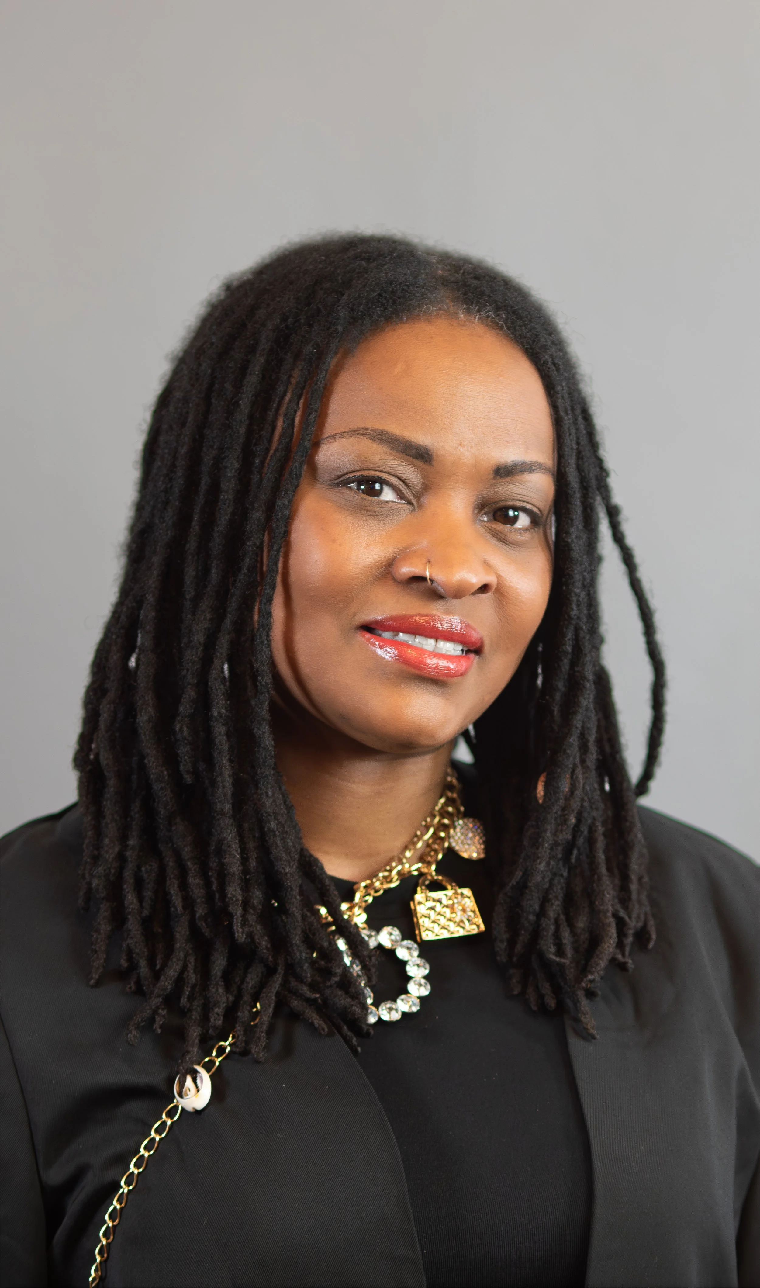 A woman with dreadlocks wearing a black top, gold jewelry, and makeup, against a plain background.