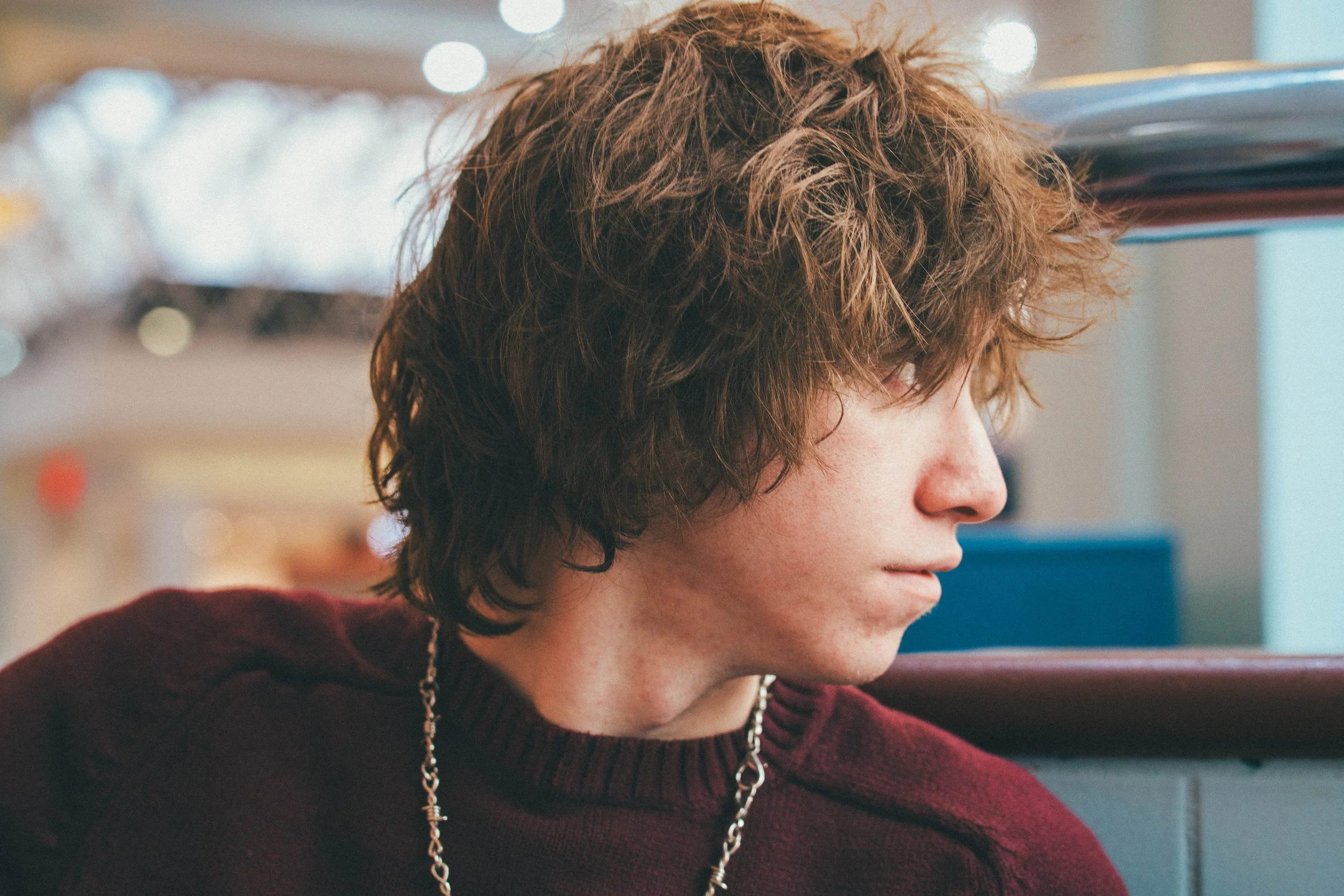 Close-up of a person with wavy, reddish-brown hair, wearing a maroon sweater and a silver chain necklace, indoors with a blurred background.