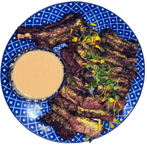 Sliced grilled beef with microgreens and a dish of creamy dipping sauce on a blue and white patterned plate.