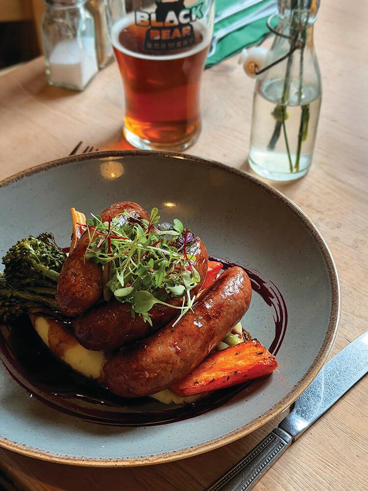 Plate of sausages on top of mash, carrots, and broccoli with gravy