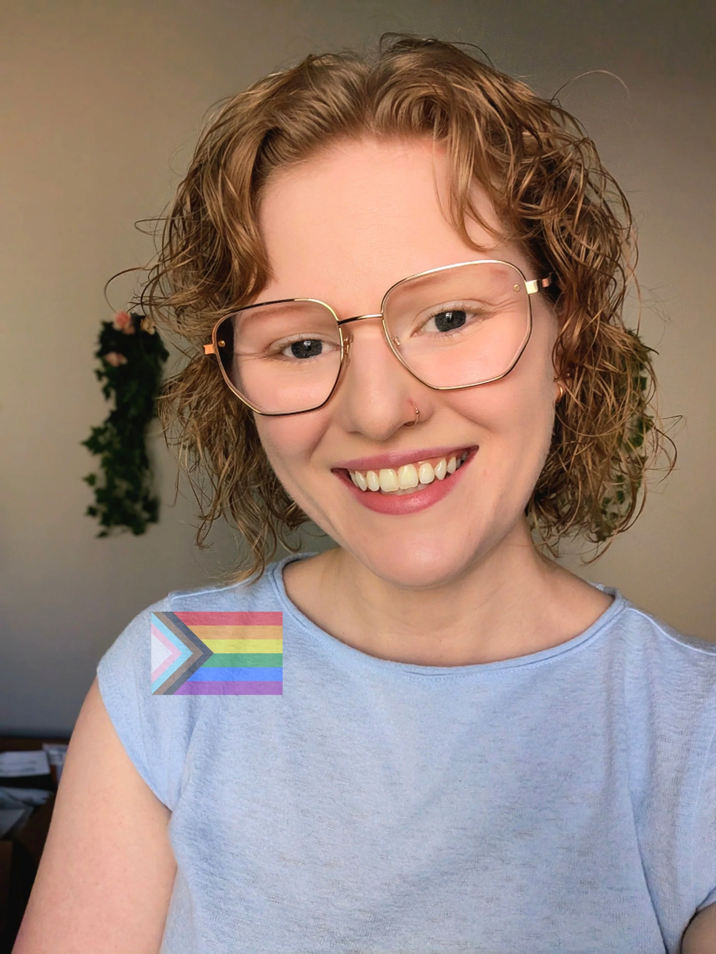 Close-up of a smiling woman with curly red hair wearing glasses and a light blue t-shirt, standing indoors with a blurred background including a couple of plants and a rainbow flag sticker.