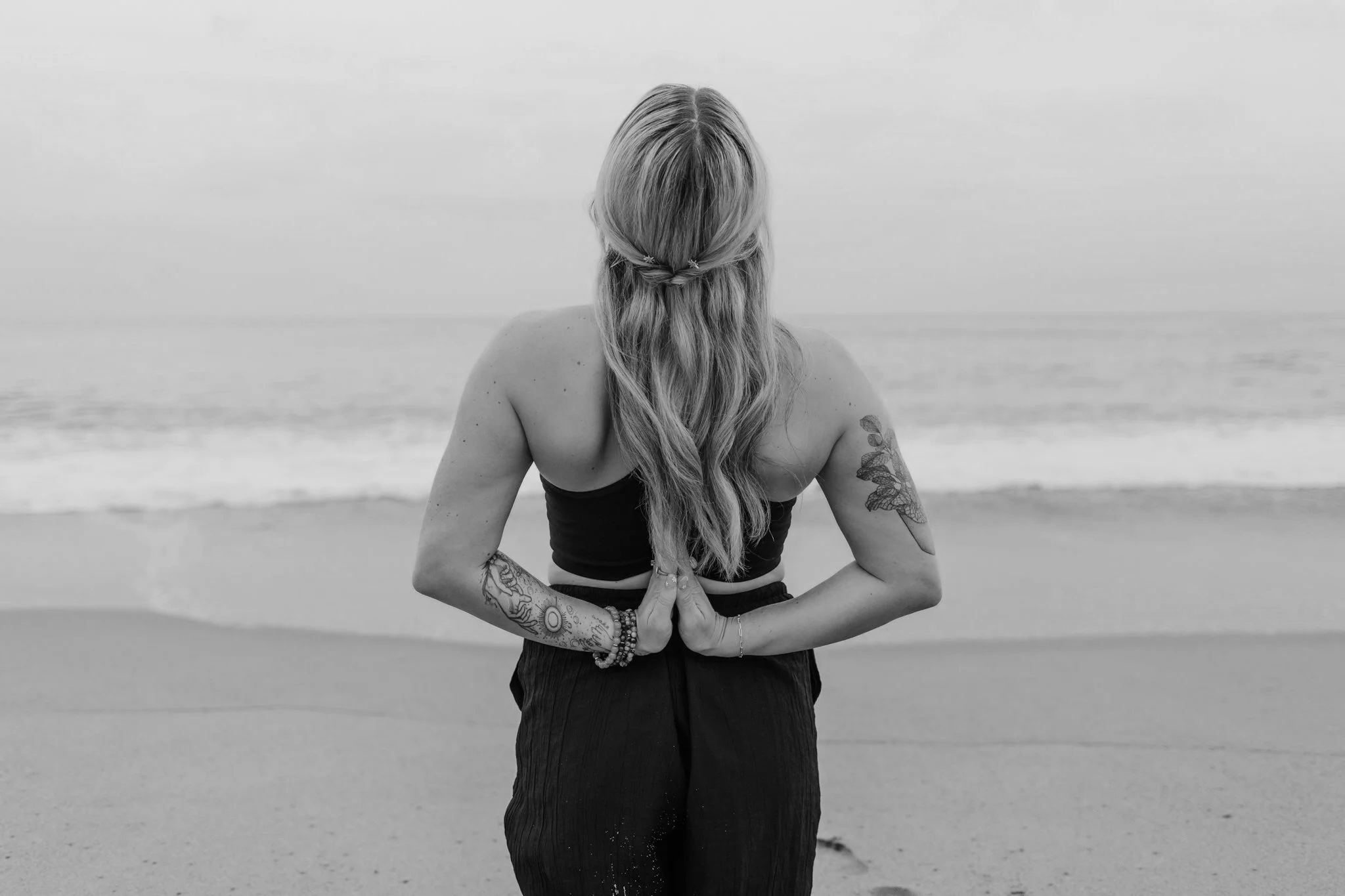 A person with long hair standing on the beach, with their back facing the camera. The person is in a yoga pose with hands pressed together in front of their chest, wearing a sleeveless top, with tattoos visible on their arms and a bracelet on their wrist.