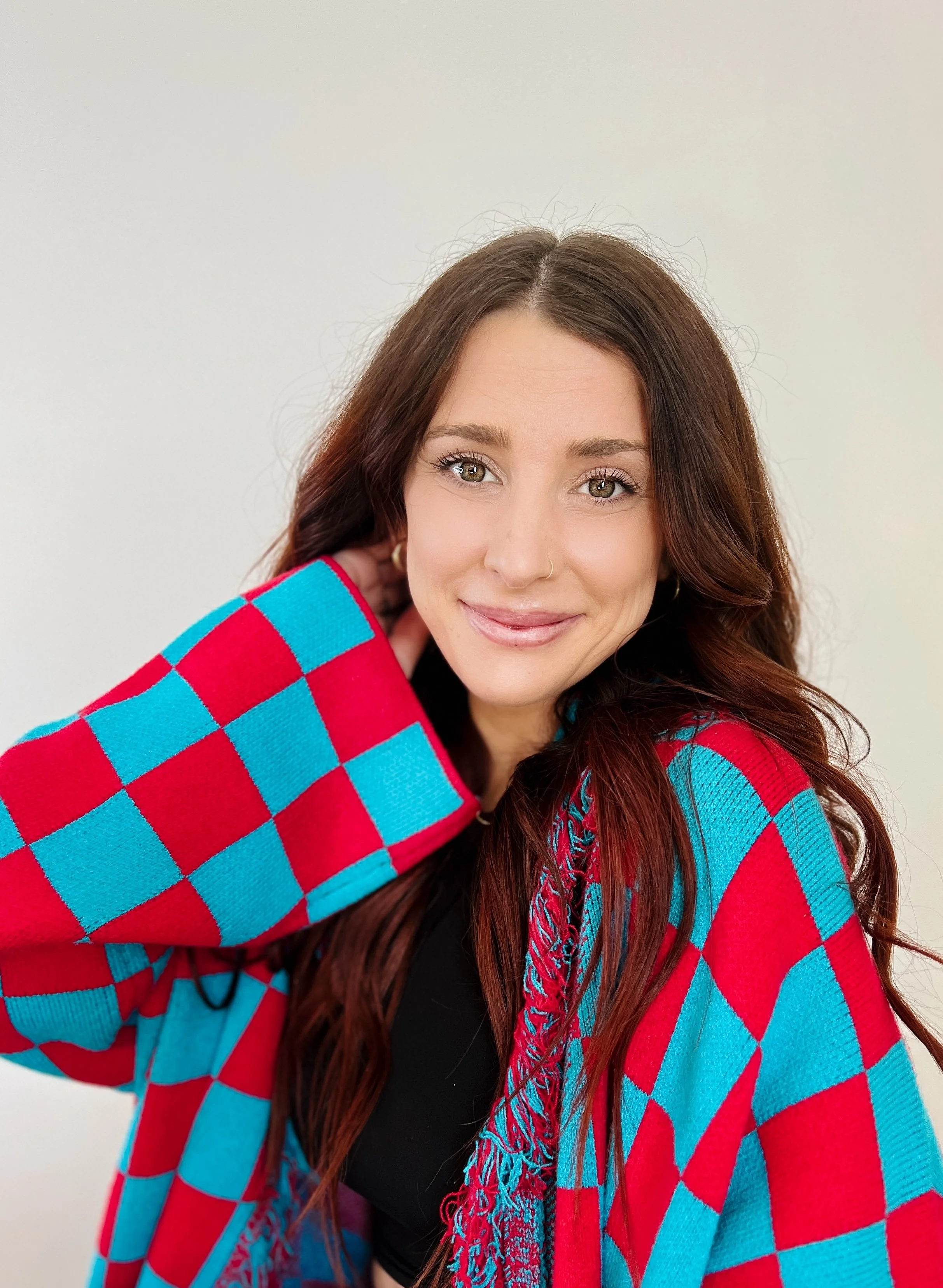 A young woman with long reddish-brown hair, hazel eyes, and a nose piercing, wearing a colorful checkered sweater with red, pink, and blue squares. She is smiling and posing with her hand behind her head against a plain light-colored background.