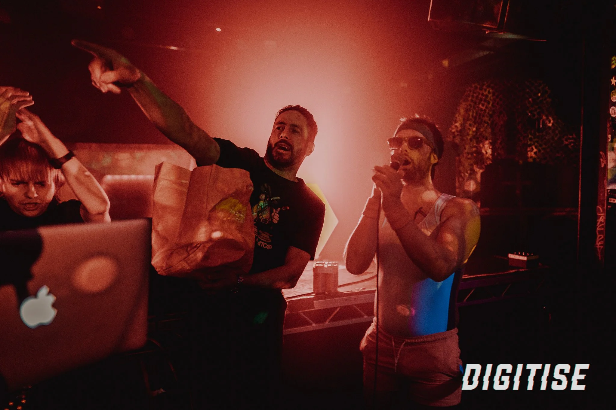Two men on a stage with a red light, one is wearing sunglasses and a tank top, holding a microphone, the other is wearing a black shirt and holding a paper bag.
