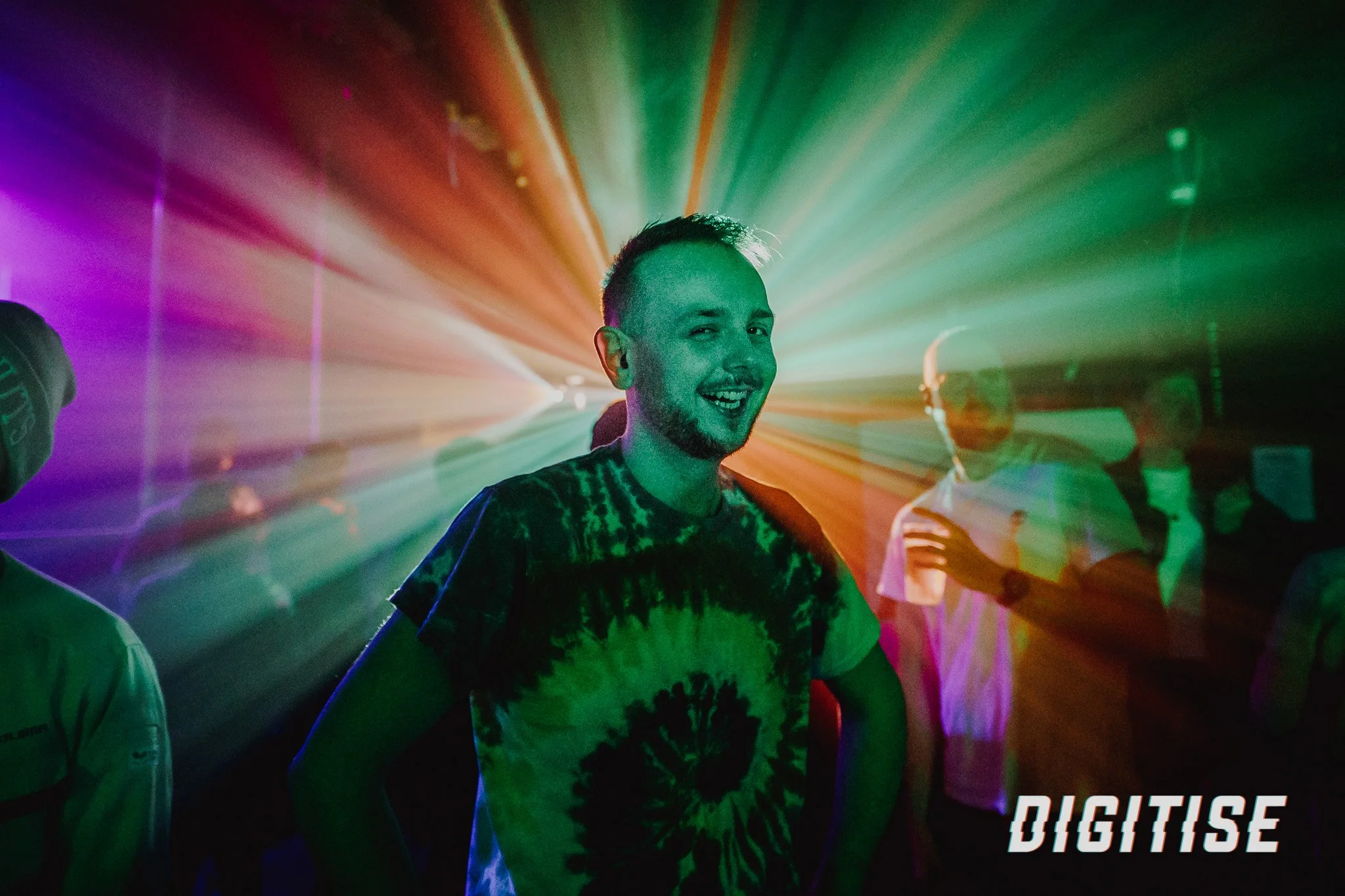 A smiling man with short hair and a beard in a nightclub with colorful laser lights.