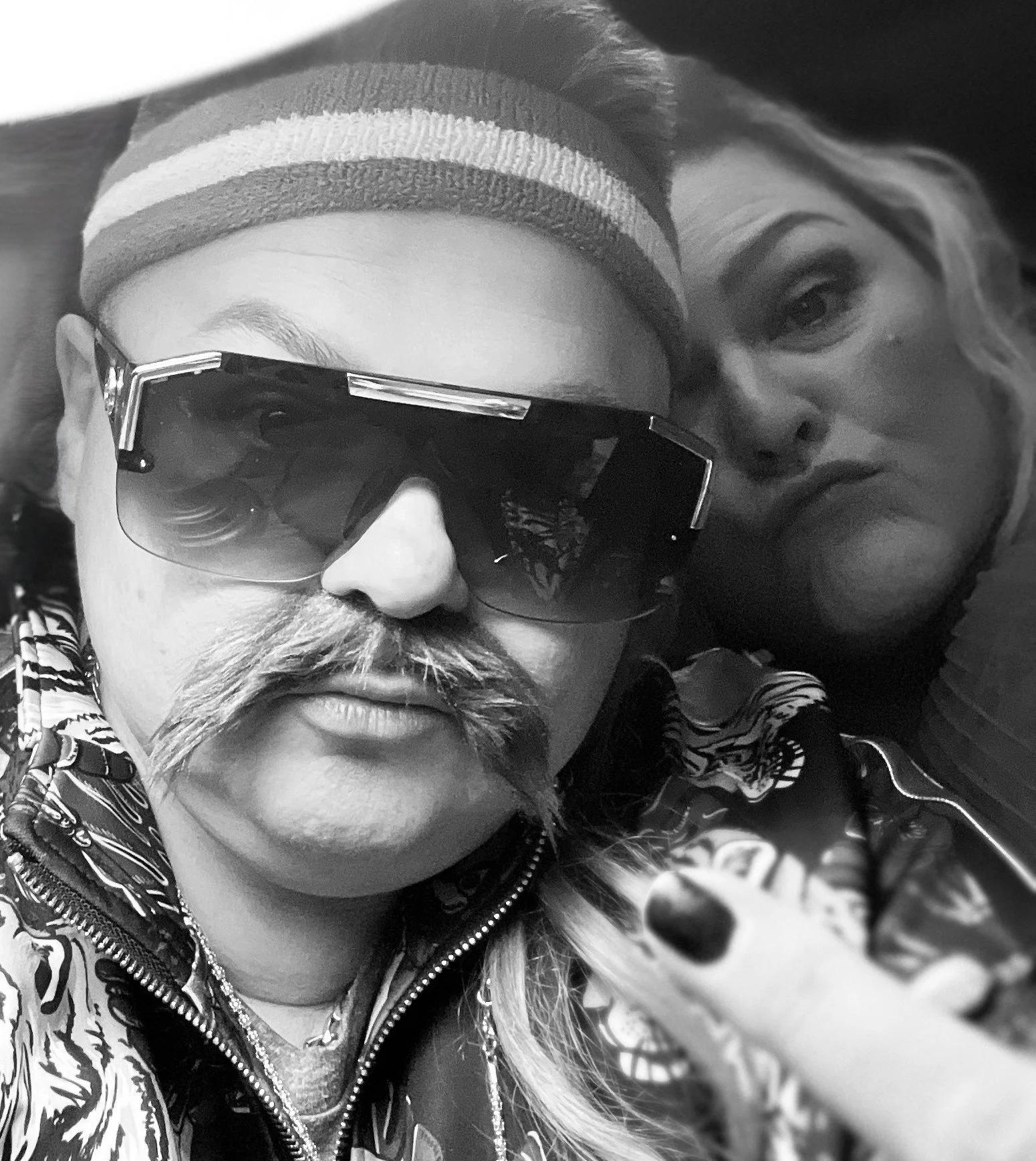 Two people taking a close-up selfie. One is wearing sunglasses, a striped beanie, and a patterned jacket. The other has long light-colored hair and is partially visible.