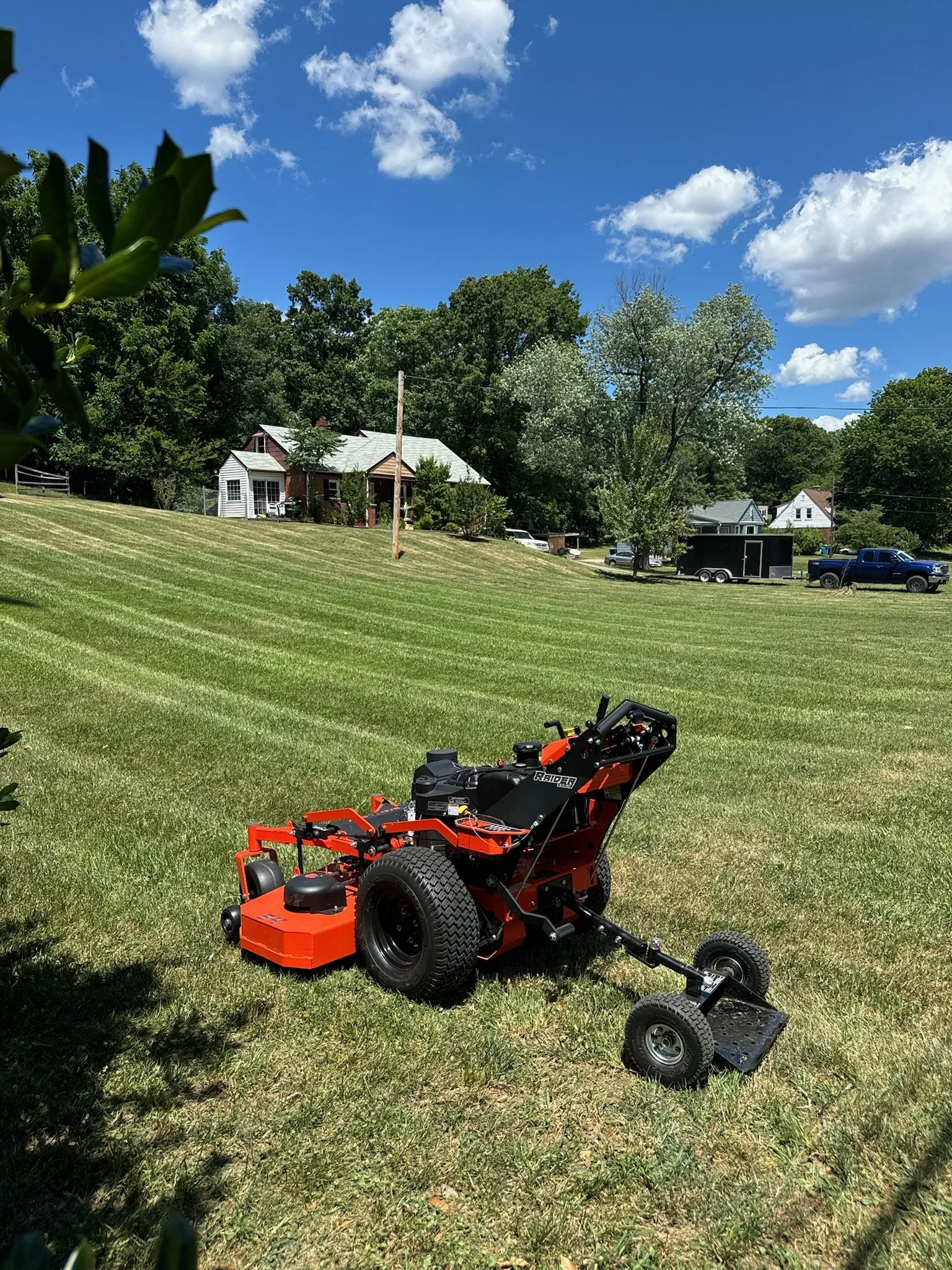A lawn tractor or mower in a green grassy yard under a bright blue sky with scattered fluffy white clouds, with houses and trees in the background.