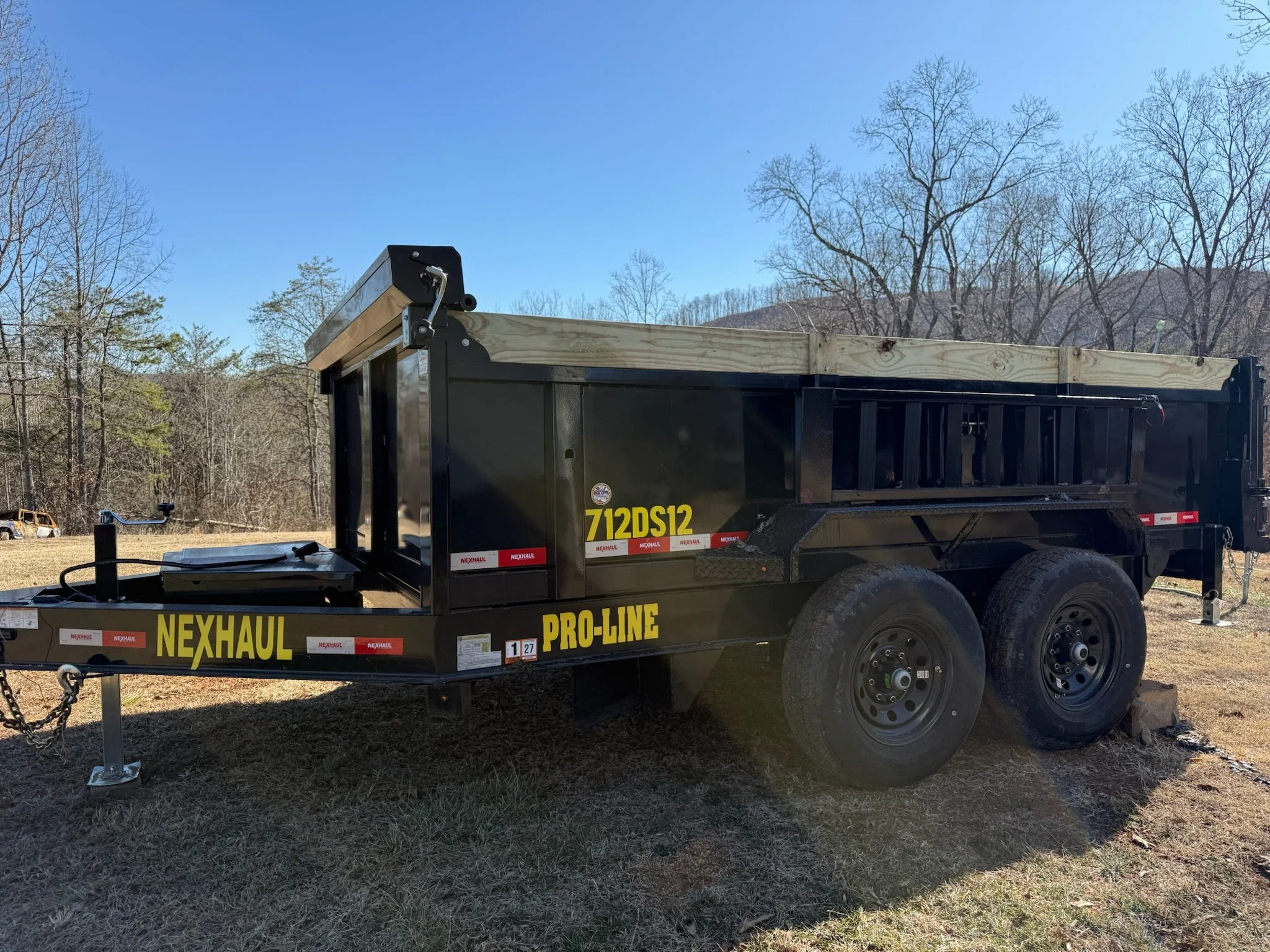 A black dump trailer with yellow and red signage, parked on grass with leafless trees and a clear blue sky in the background.
