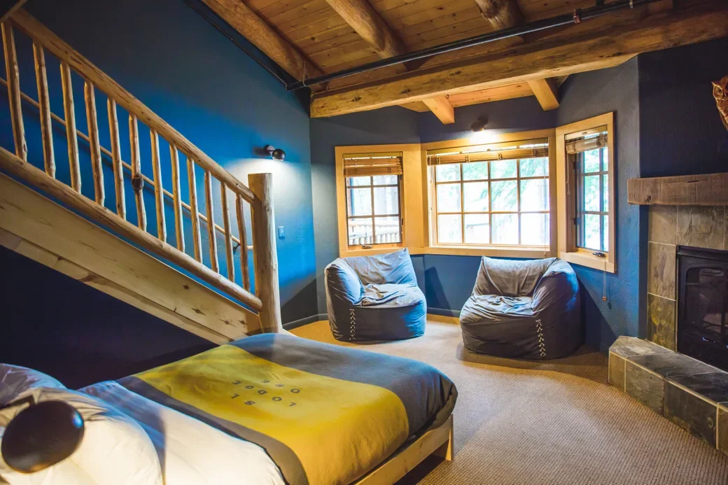 A cozy room with wooden ceiling beams, blue walls, and large windows with blinds. There are two bean bags near the windows and a bed with a yellow and black comforter.