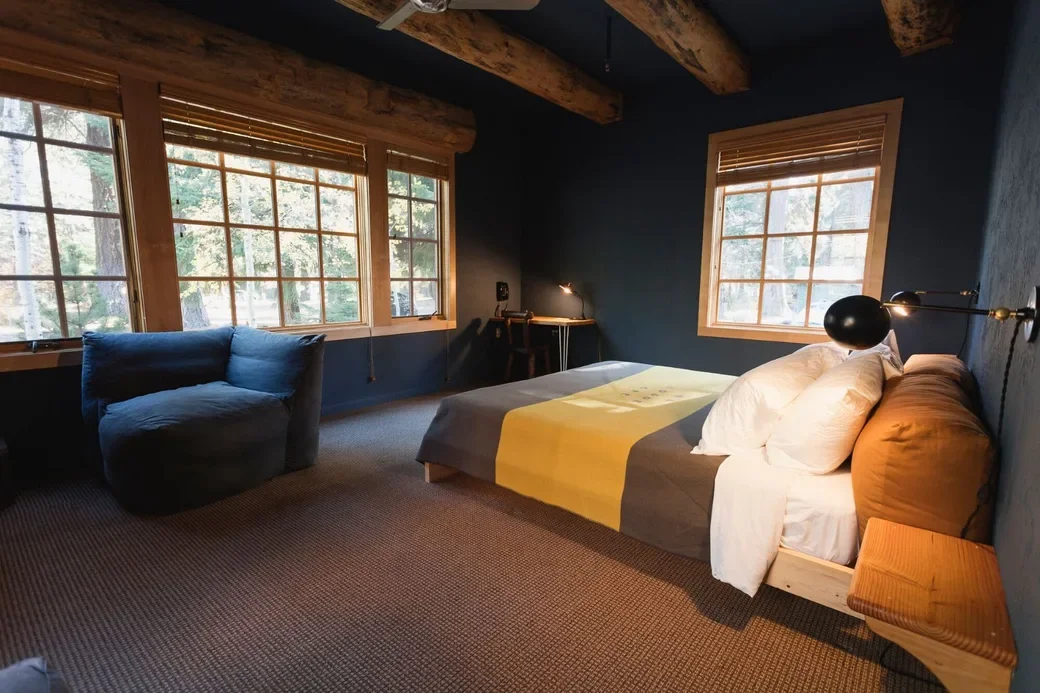 Cozy bedroom with a wooden ceiling, large windows with blinds, a bed with white pillows and a multicolored duvet, a black armchair, and a small desk with a lamp.