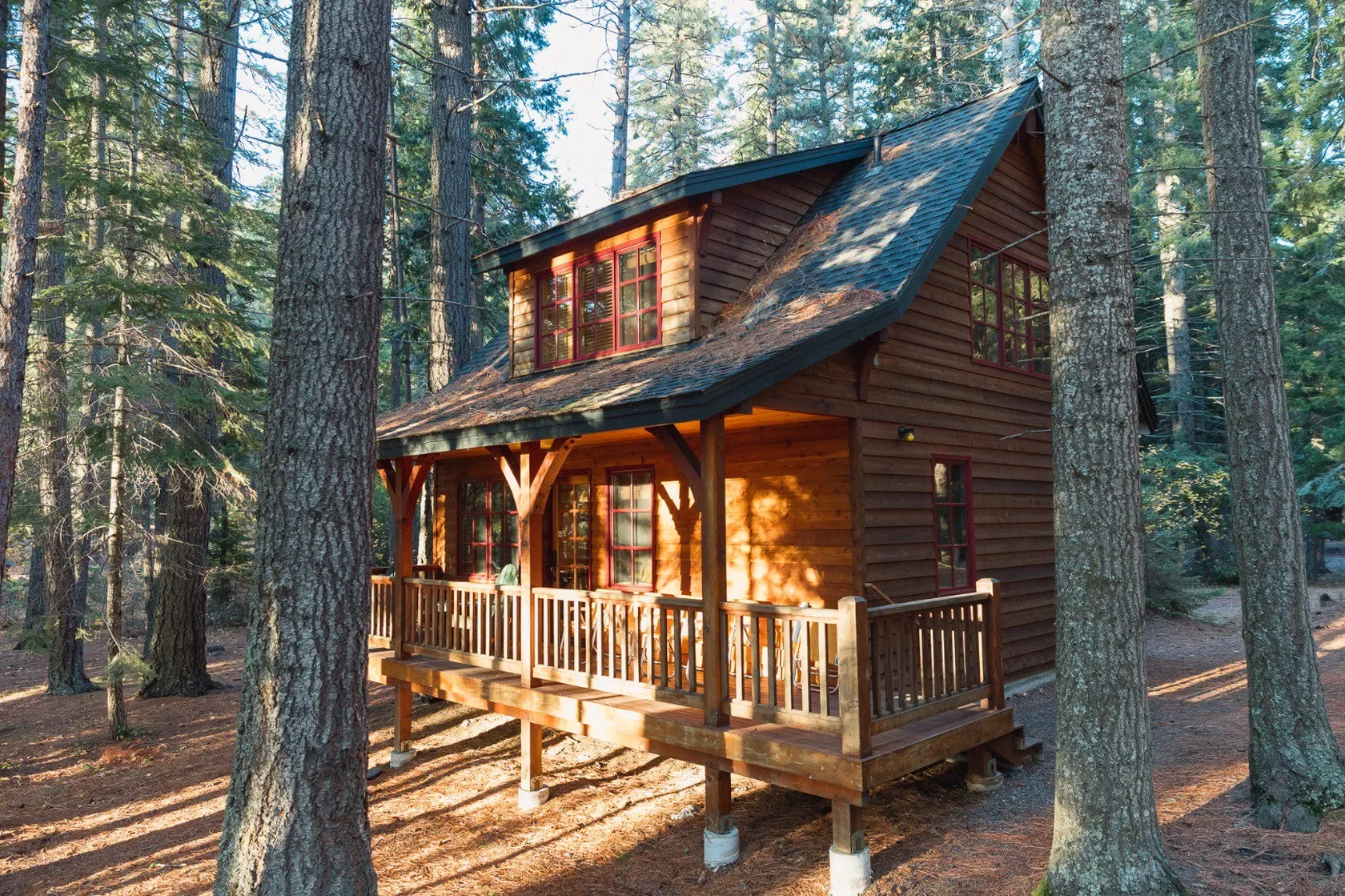 A two-story wooden cabin with a porch, set among tall pine trees in a forest.